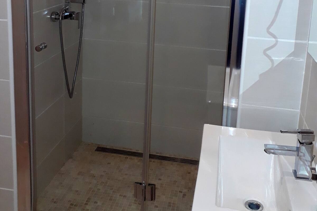 Eine gläserne Duschkabine, ein weißes Handtuch hängt an der Tür, ein Waschbecken mit Spiegel und geflieste Wände.
