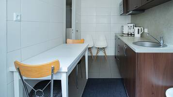 A compact kitchen with a white countertop, a sink, and a microwave. There are two white chairs and a wooden chair.