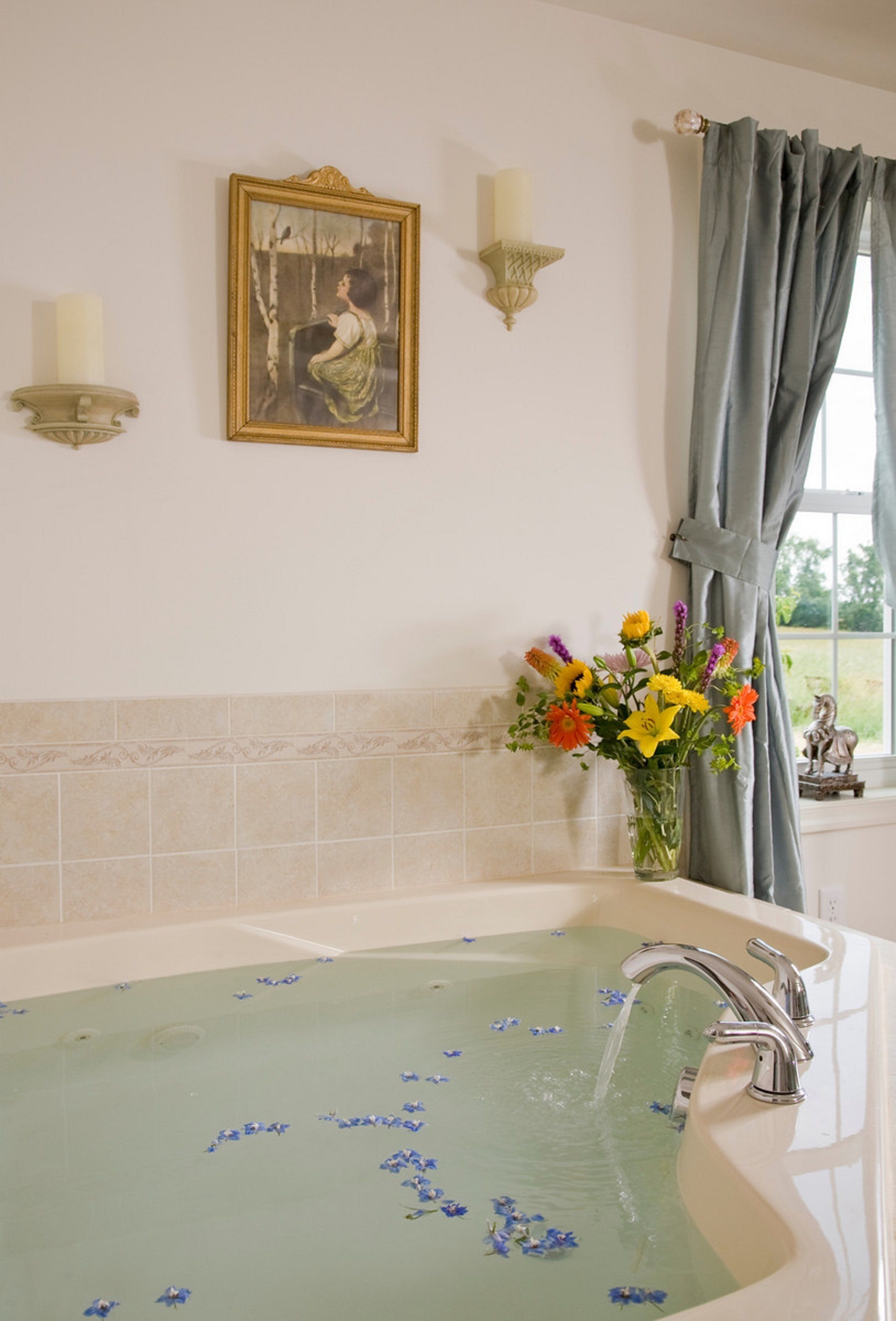Un baño con bañera, un jarrón de flores, una lámpara de pared y un cuadro enmarcado.