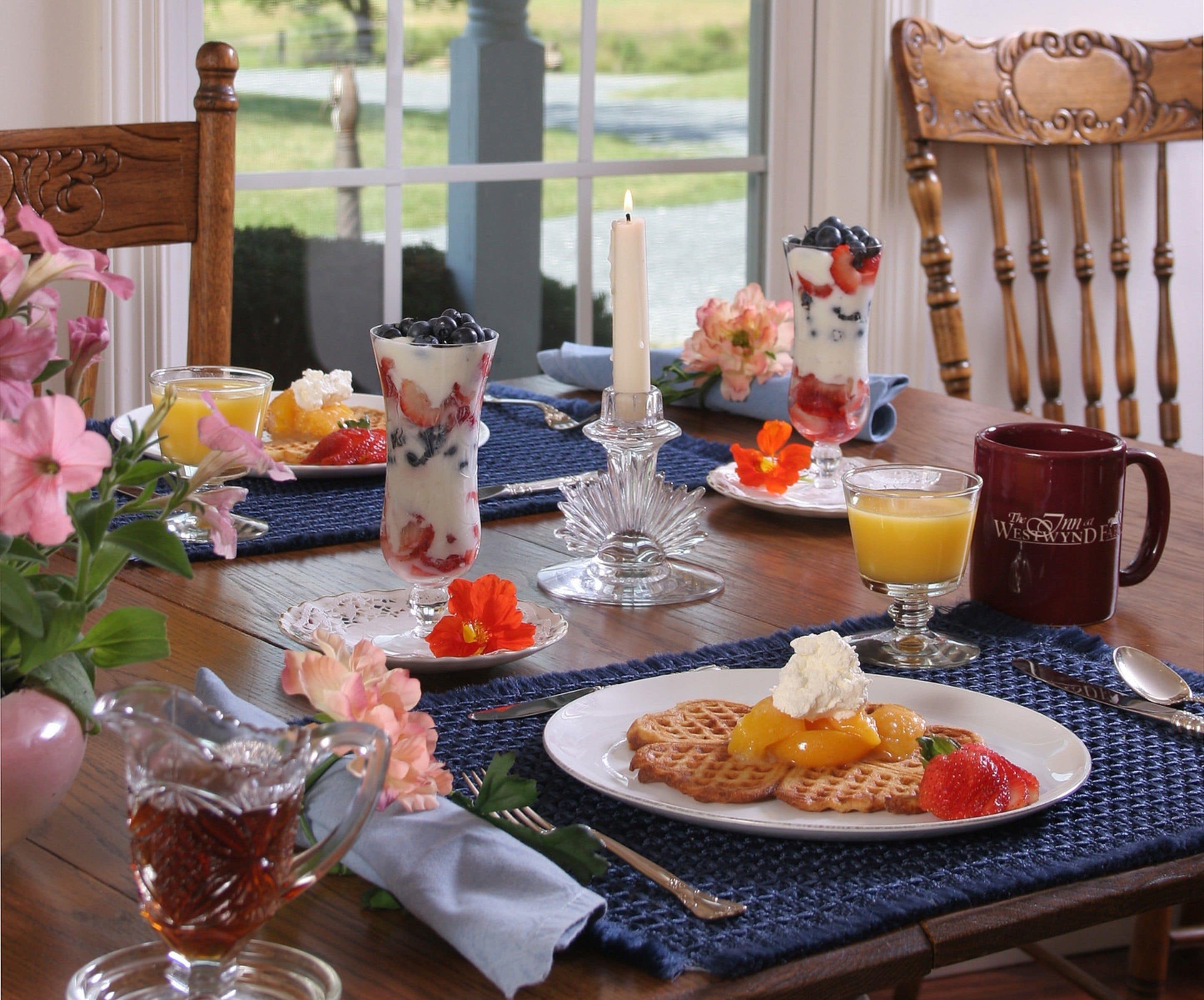 Une table mise pour le petit-déjeuner, avec une nappe pour deux personnes, comprenant des gaufres, des fruits, du yaourt et une tasse de café.