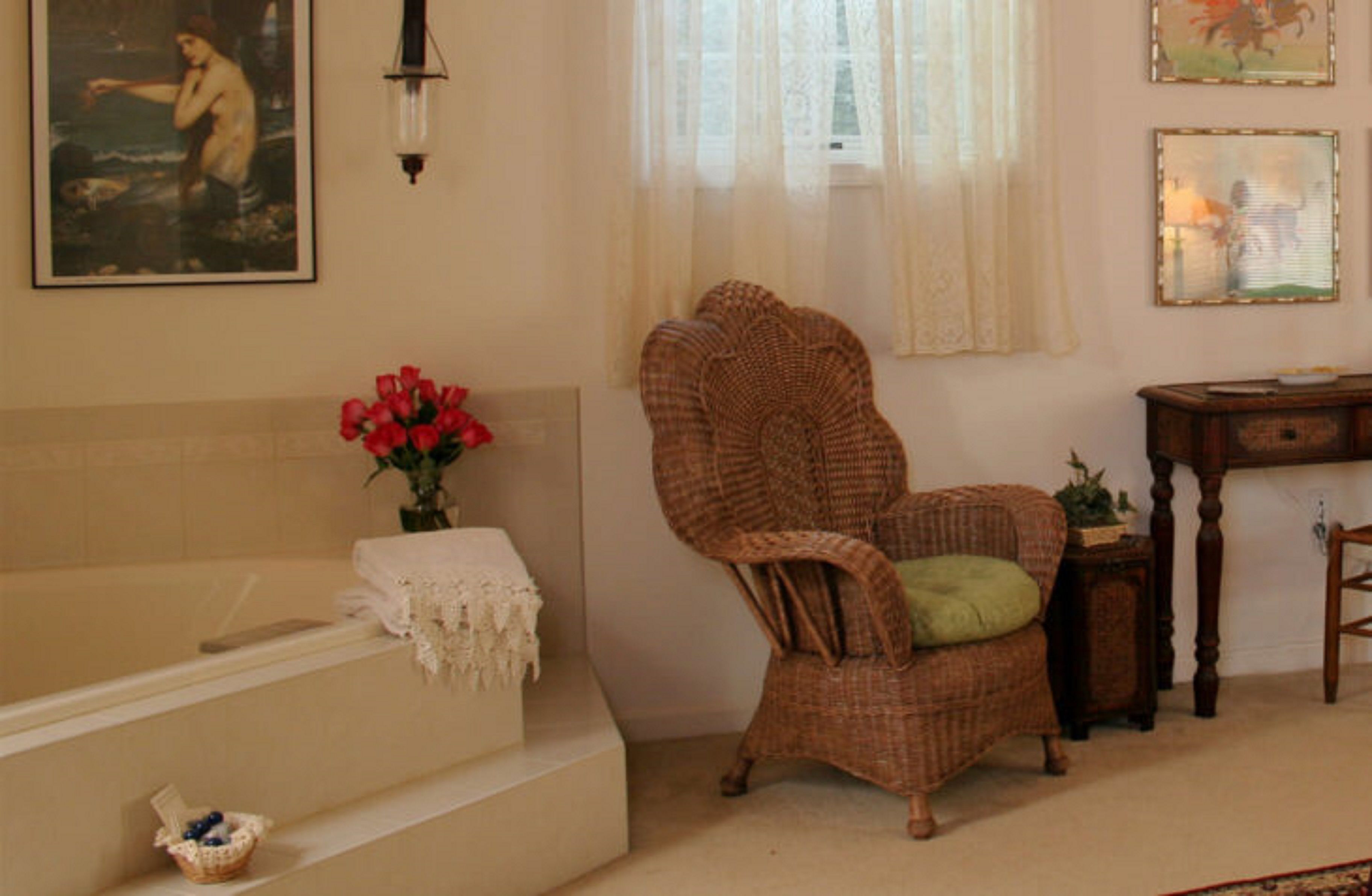 A room with a bathtub, wicker chair, and a small table with a lamp.