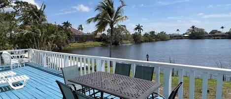 A blue deck with a table and chairs, overlooking a calm lake with palm trees and houses in the background.
