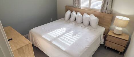 A neatly made bed with white bedding and a wooden headboard, a bedside table with a lamp, and a window with curtains.