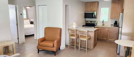 A modern hotel room with a bed, a sitting area with a chair, a small table, and a kitchenette with a microwave, stove, and cabinets.