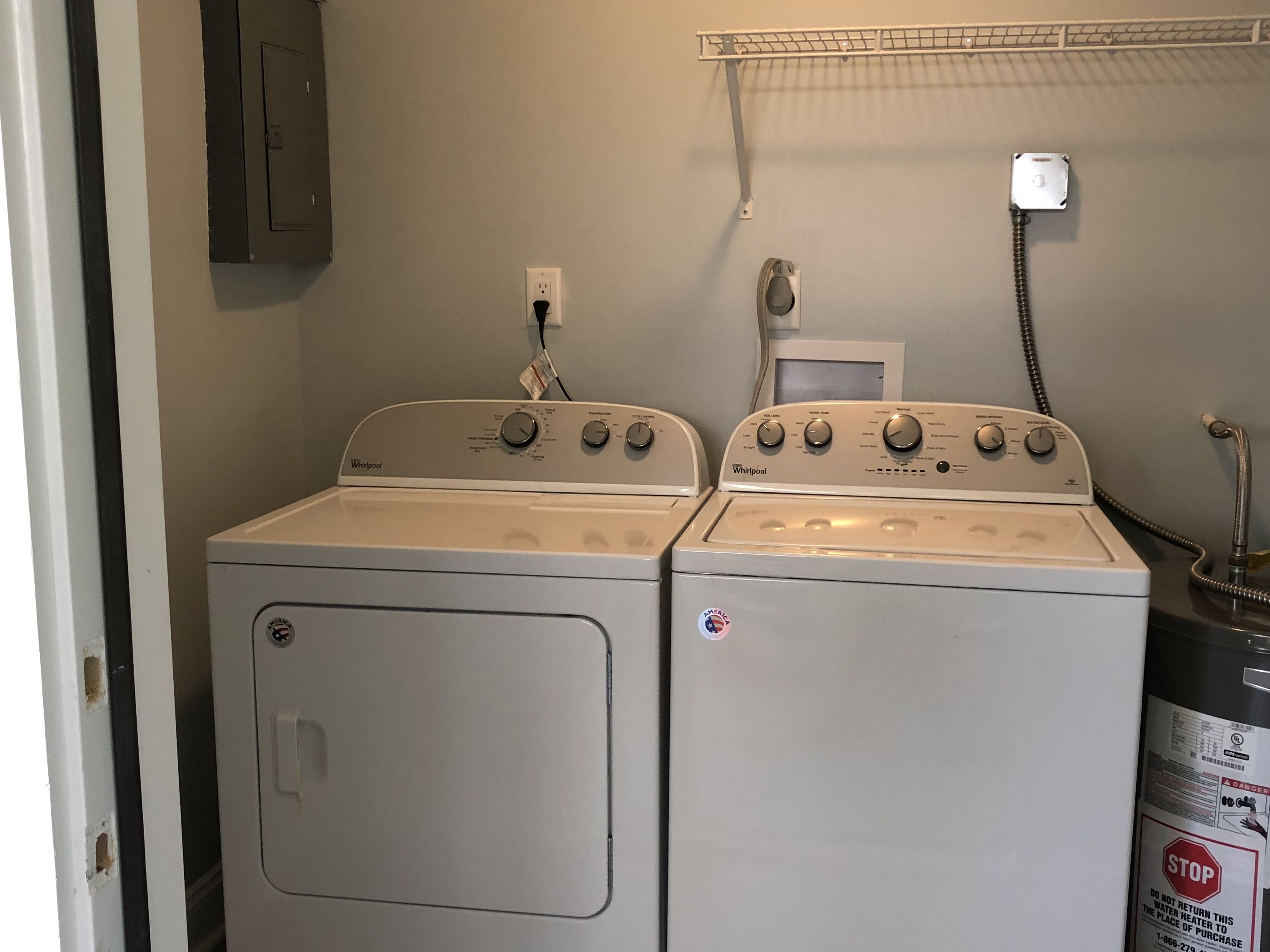 Pesuhuoneessa on kaksi Whirlpool-merkkiä olevaa pesu- ja kuivauskonetta, lämminvesivaraaja ja seinällä metallinen teline.