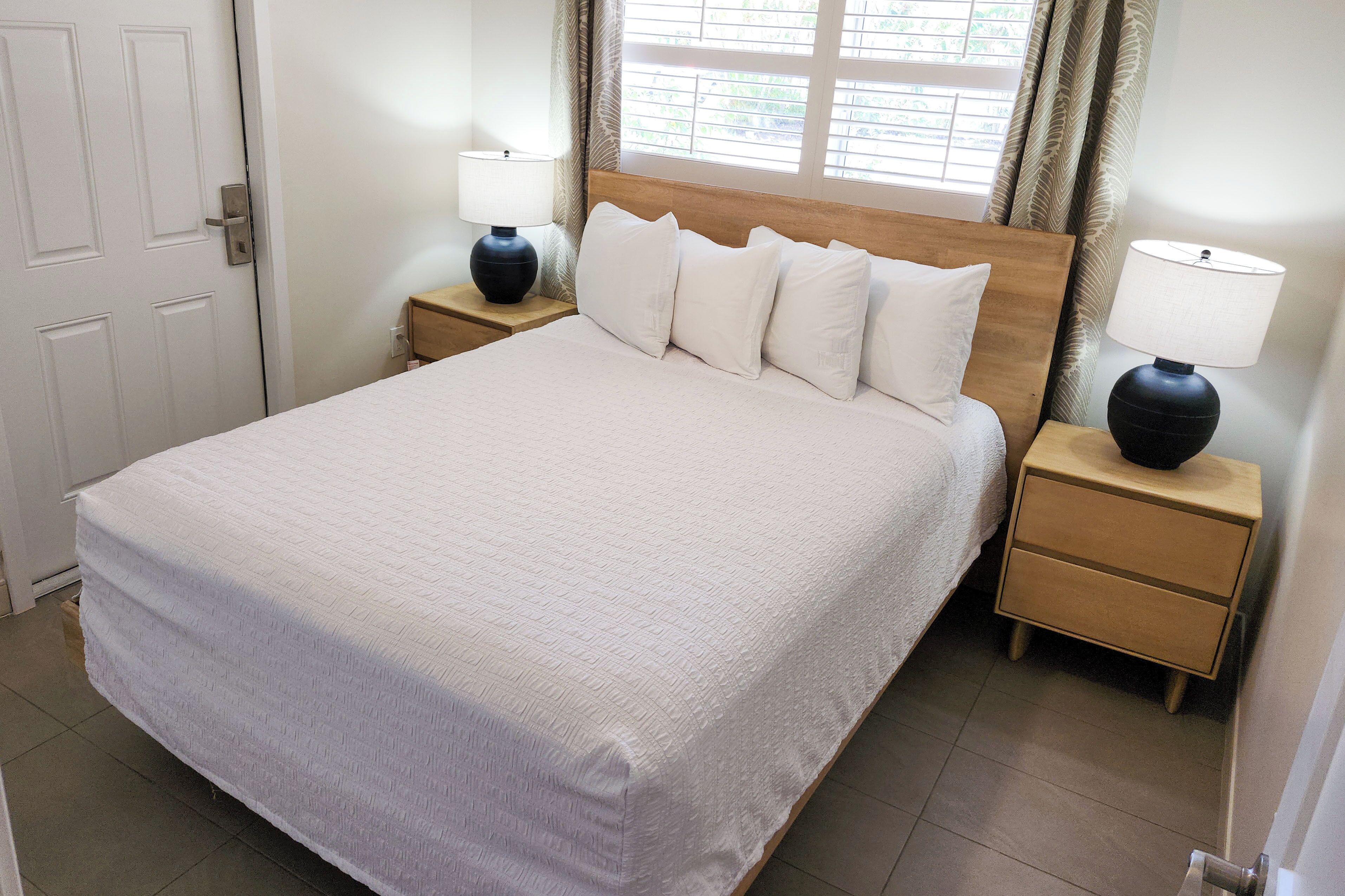 A neatly made bed with a white bedspread and pillows, two bedside tables with lamps, a wooden headboard, and a window with curtains.