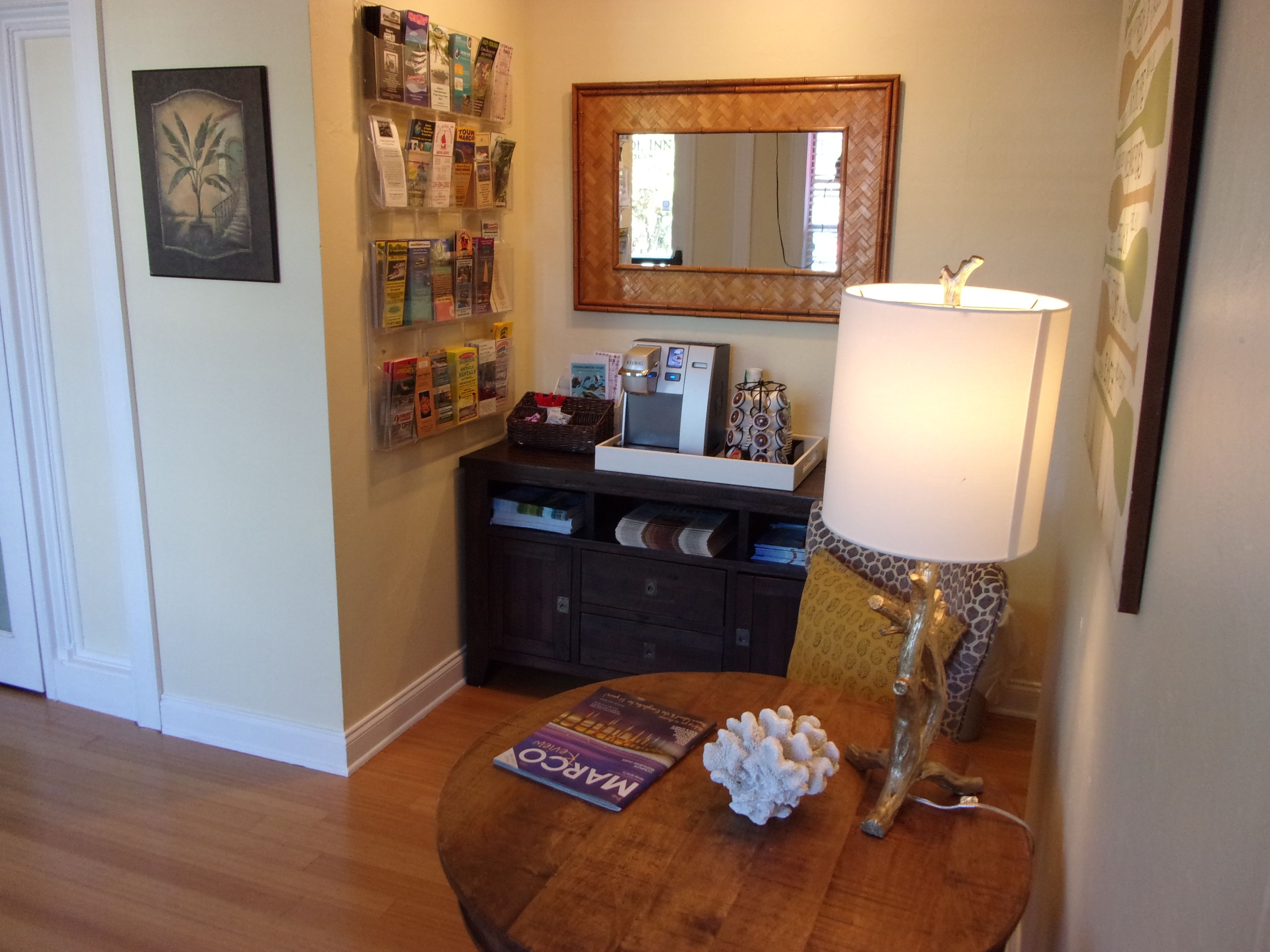 A room with a wooden table, a lamp, a mirror, a bulletin board with various notices, and a framed picture on the wall.
