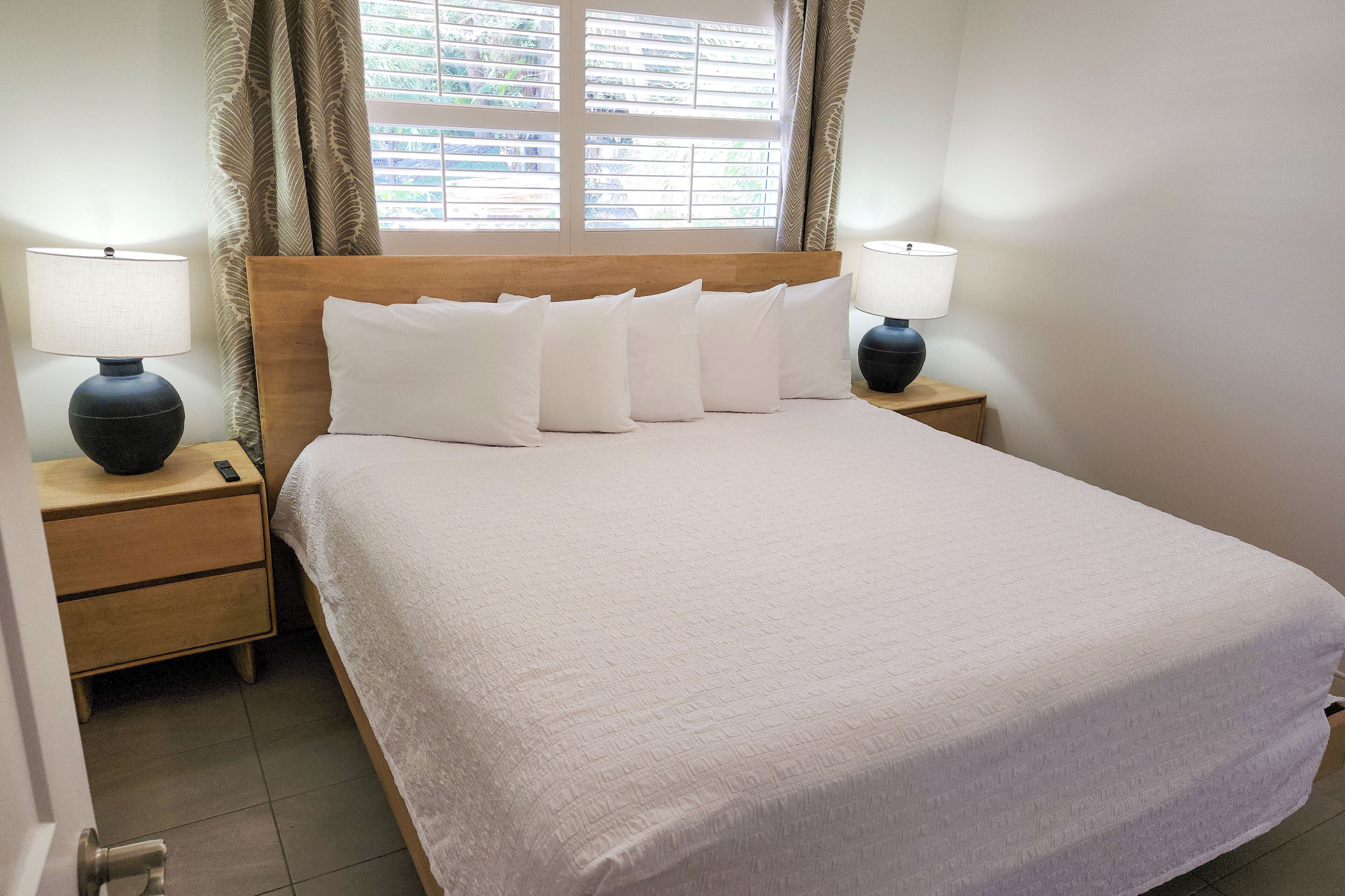 Una cama bien hecha con sábanas y almohadas blancas, dos lámparas de noche y una ventana con cortinas.