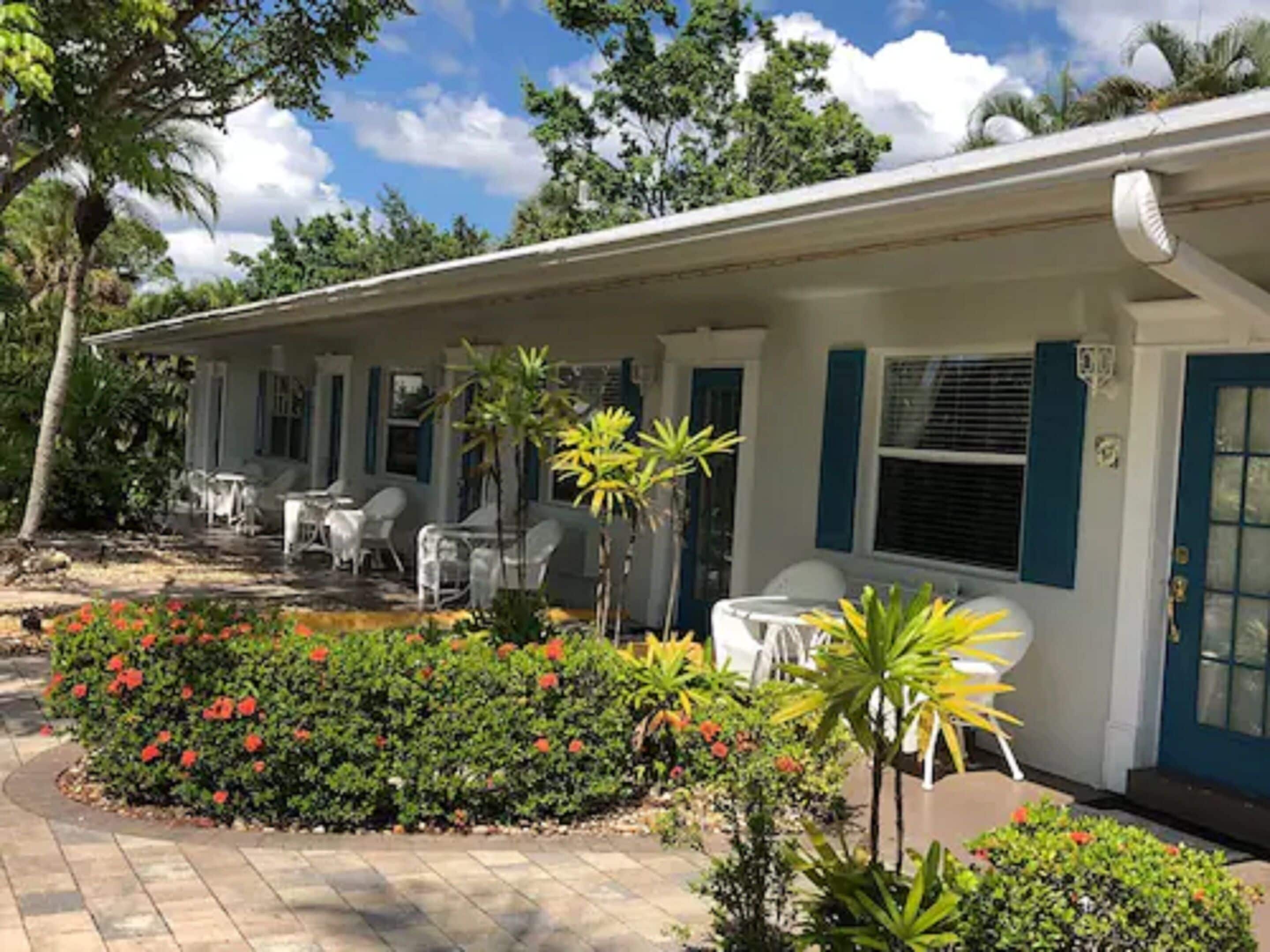 A single-story motel with a covered patio area, outdoor seating, and a well-maintained garden with red flowers and tropical plants.