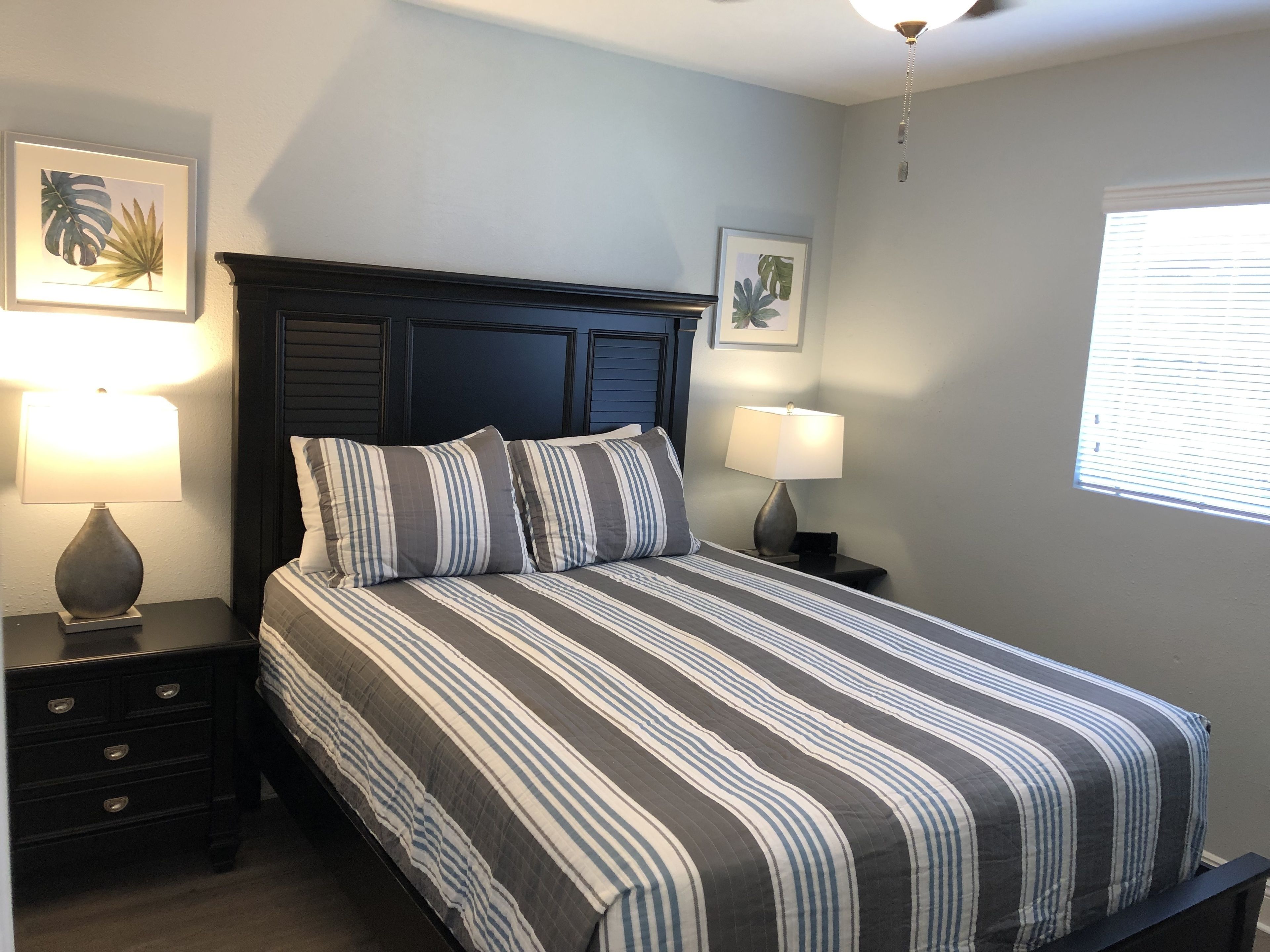 A bedroom with a dark wood headboard, striped bedding, two bedside tables with lamps, and framed tropical leaf artwork on the walls.