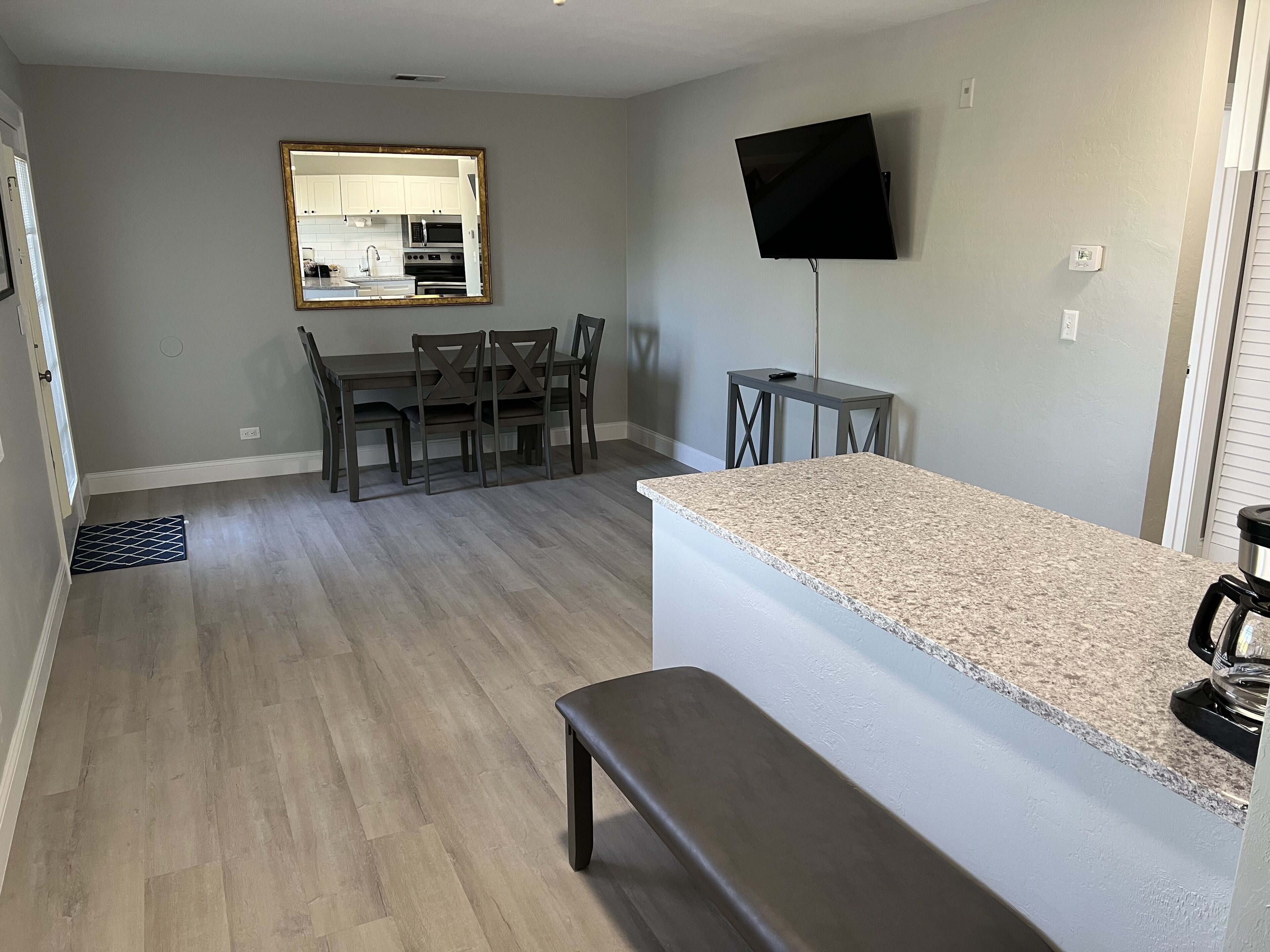 A modern kitchen with a granite countertop, a dining area with a table and chairs, a large mirror, and a flat-screen TV mounted on the wall.