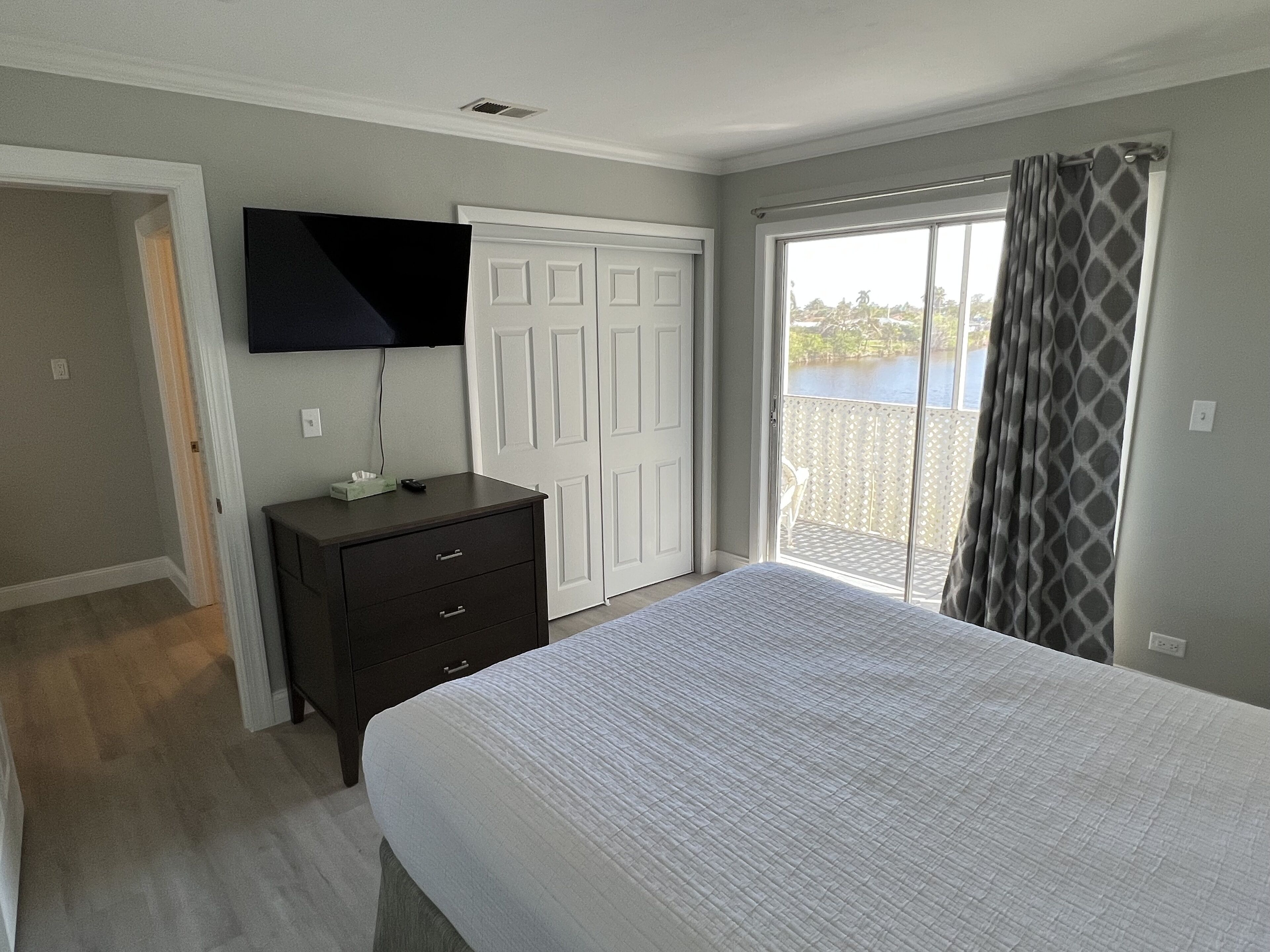 Quarto com cama, cómoda, televisão, porta de correr para uma varanda e porta branca.