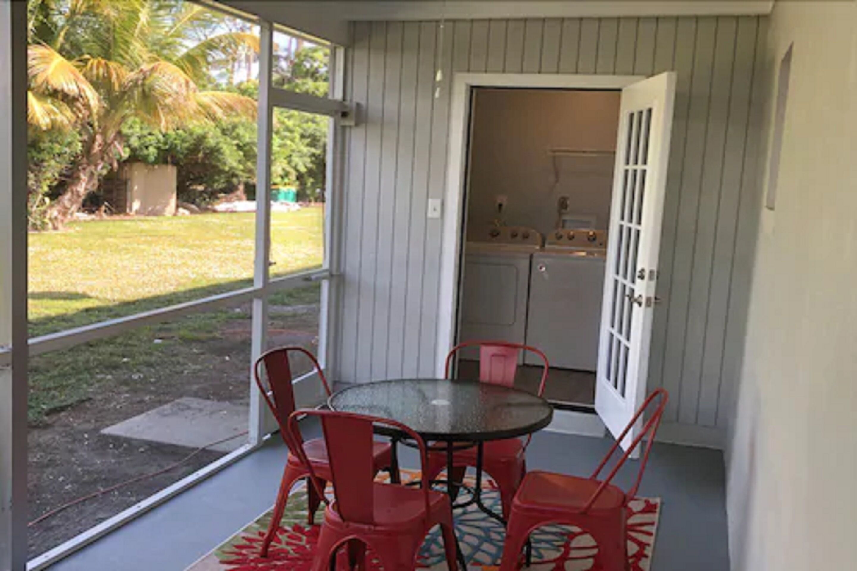 Un patio cubierto con una mesa redonda y sillas rojas, una puerta que da a una cocina, y vistas a un césped y una palmera.