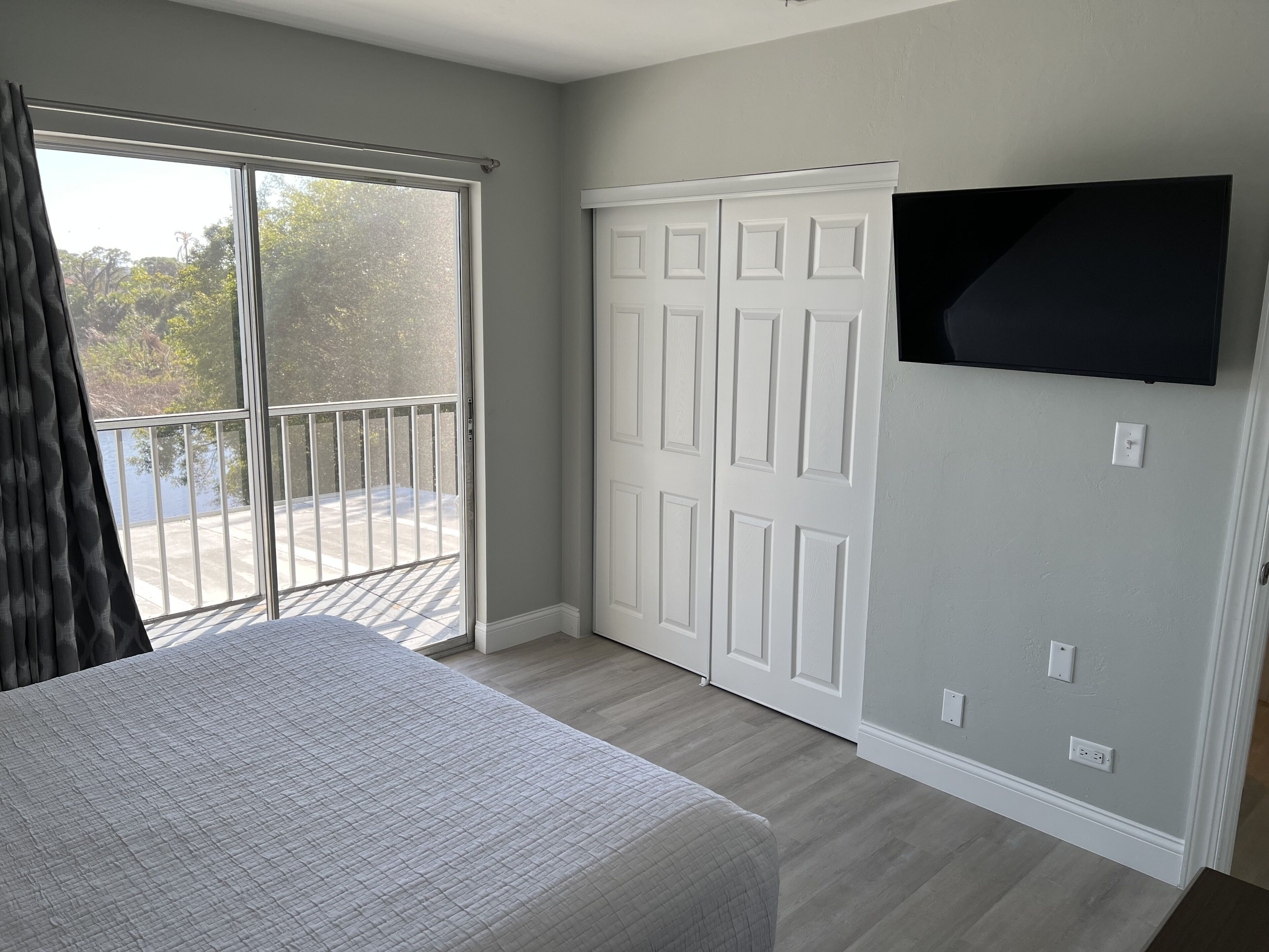 Un dormitorio con cama, una puerta corrediza de vidrio a un balcón, un armario blanco y un televisor de pared.