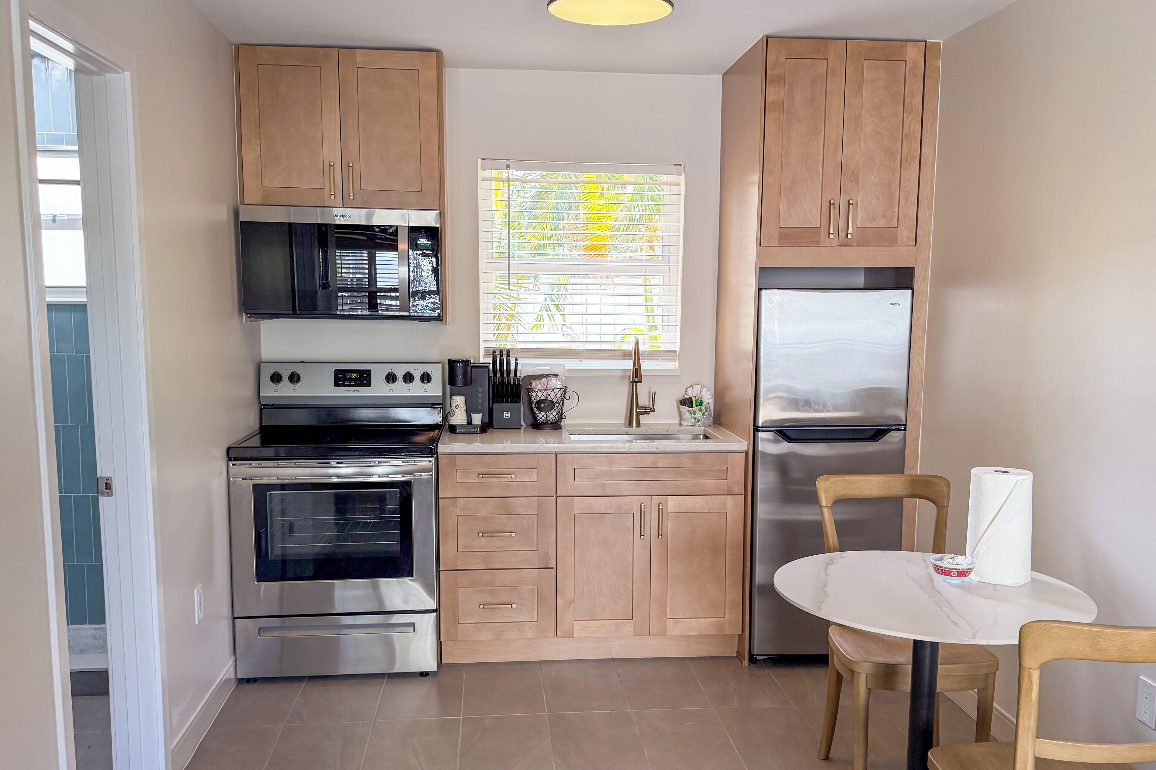 A compact kitchen with stainless steel appliances, wooden cabinets, a window with blinds, and a small dining area.