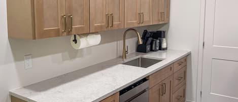 A kitchen with wooden cabinets, a stainless steel dishwasher, a marble countertop, and a sink with a gold faucet.