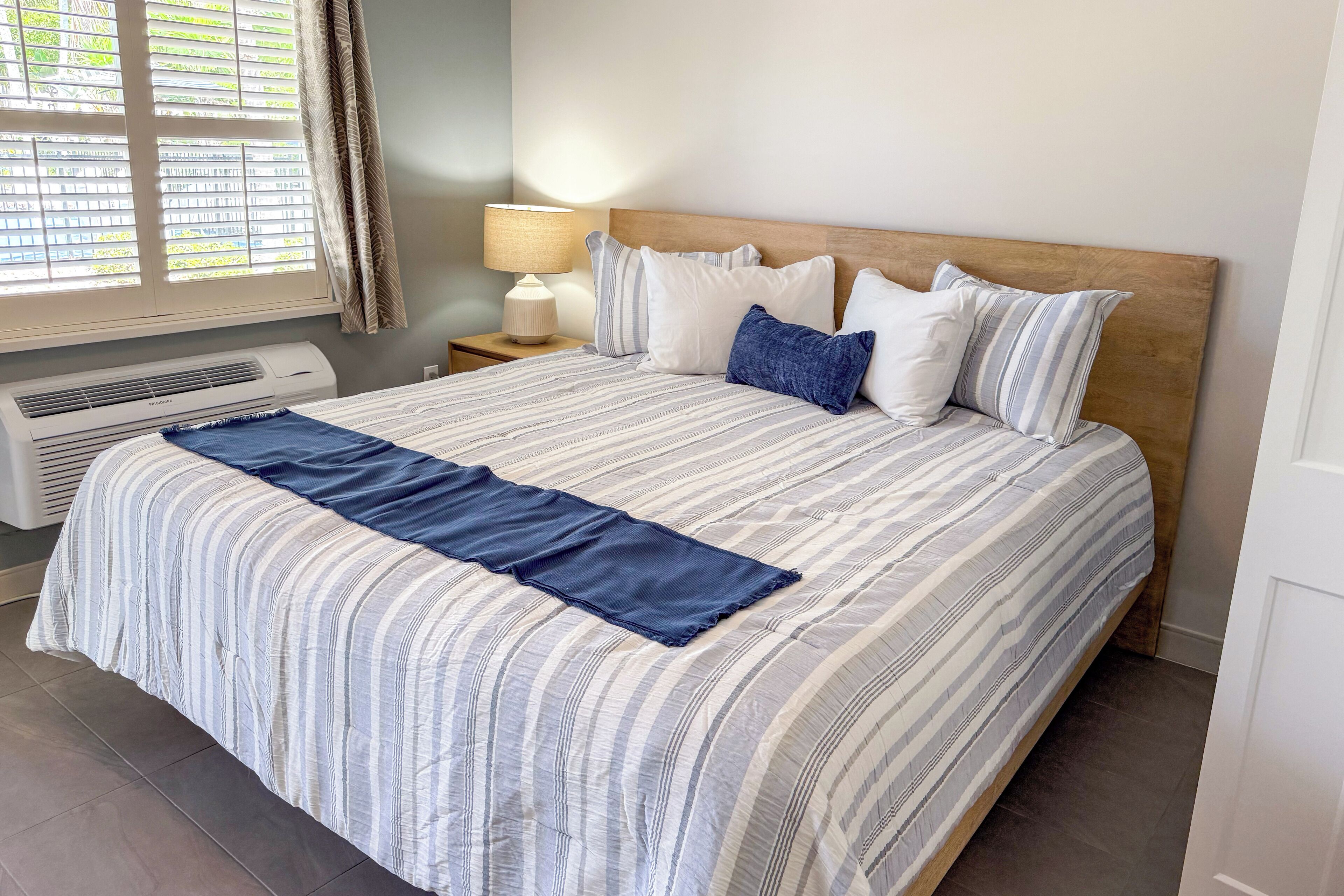 A neatly made bed with a striped bedspread and pillows, a bedside lamp, and an air conditioning unit.