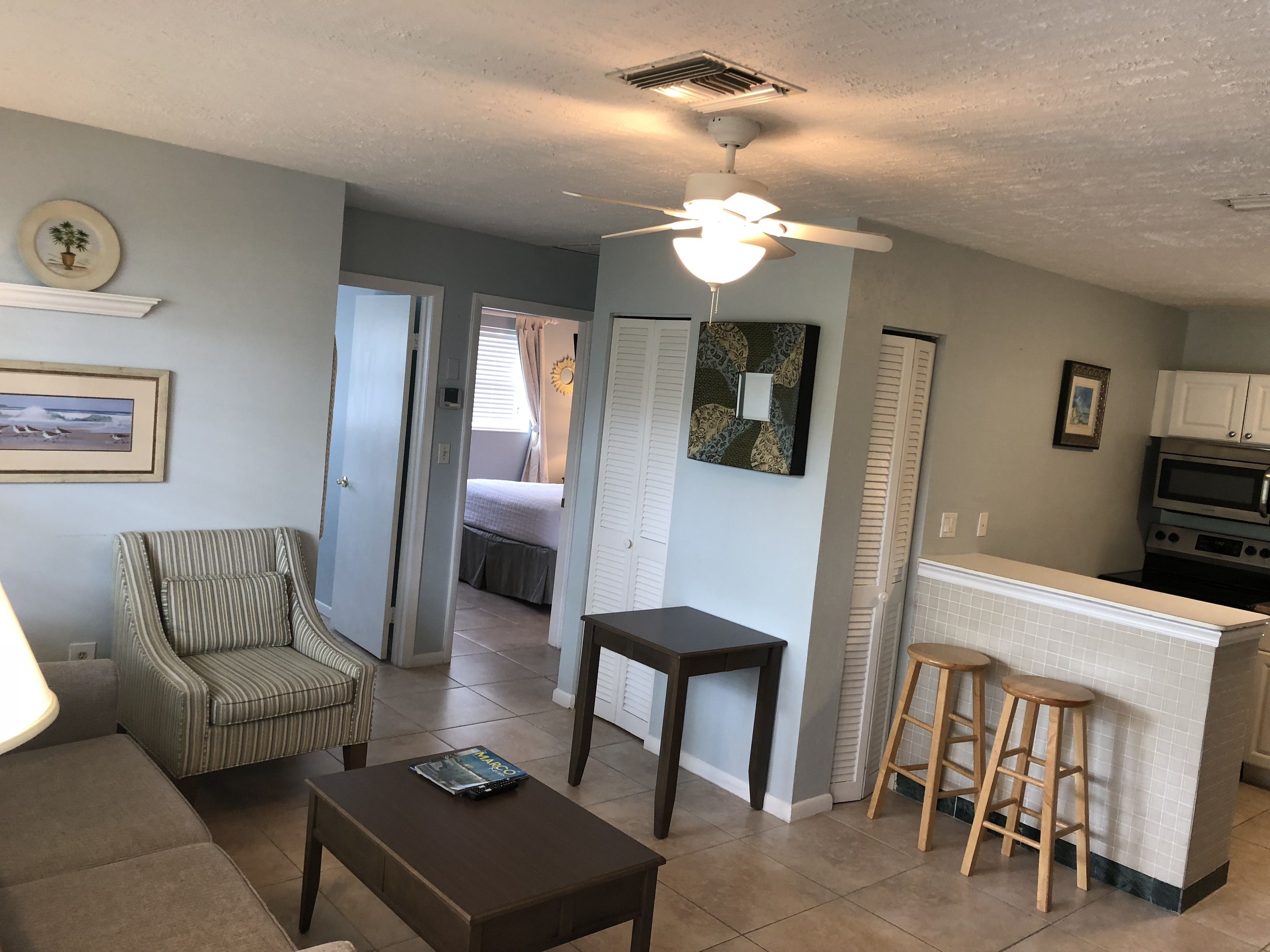 A living room with a sofa, a coffee table, a striped armchair, a small table, and a ceiling fan. There is a kitchen area with a microwave, a stove, and a bar stool.