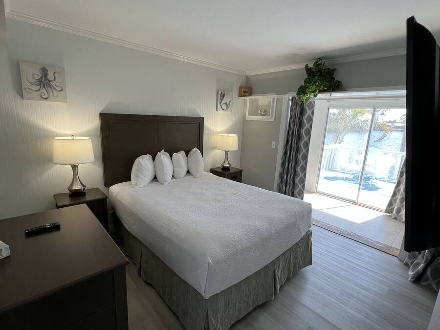 A hotel room with a large bed, two bedside tables with lamps, a remote control on a side table, and a view of the outdoors through a sliding door.