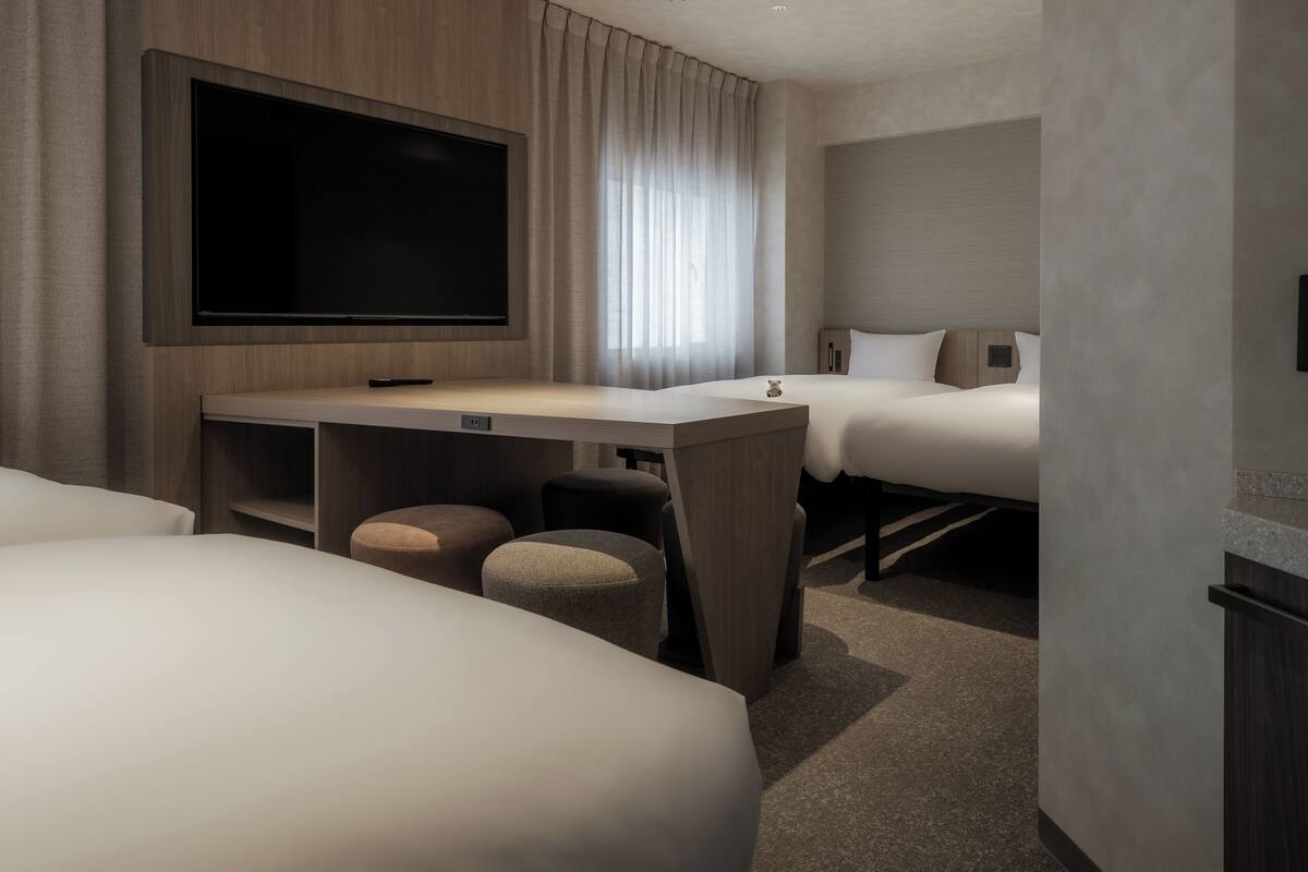 A hotel room with a large flat-screen TV mounted on the wall, a desk with a chair, two beds, and a small ottoman.