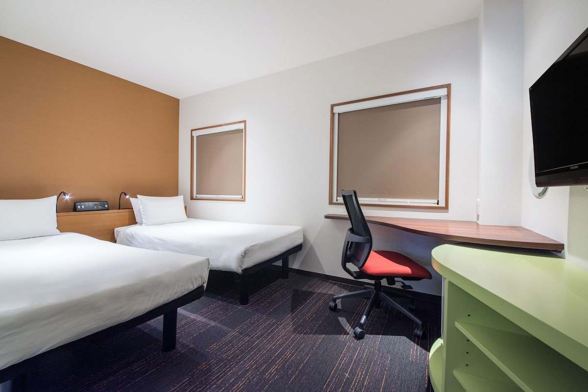 A hotel room with two single beds, a desk with a chair, a television, and a green side table.