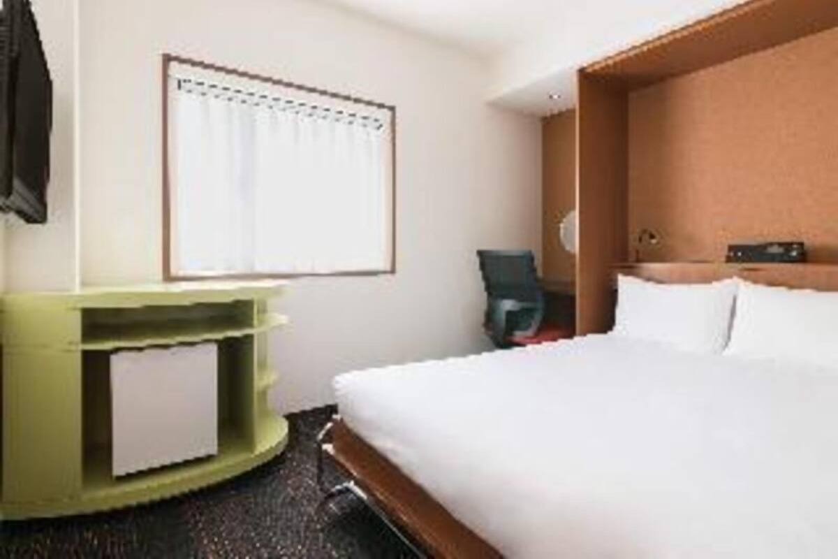 A hotel room with a bed, a desk, a chair, a window with curtains, and a wall-mounted clock.