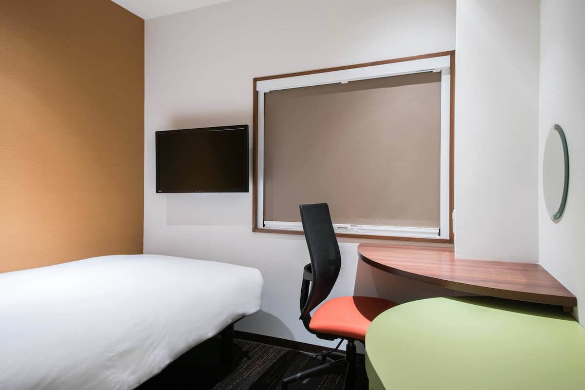 A hotel room with a single bed, a desk with a chair, a flat-screen TV mounted on the wall, and a window.