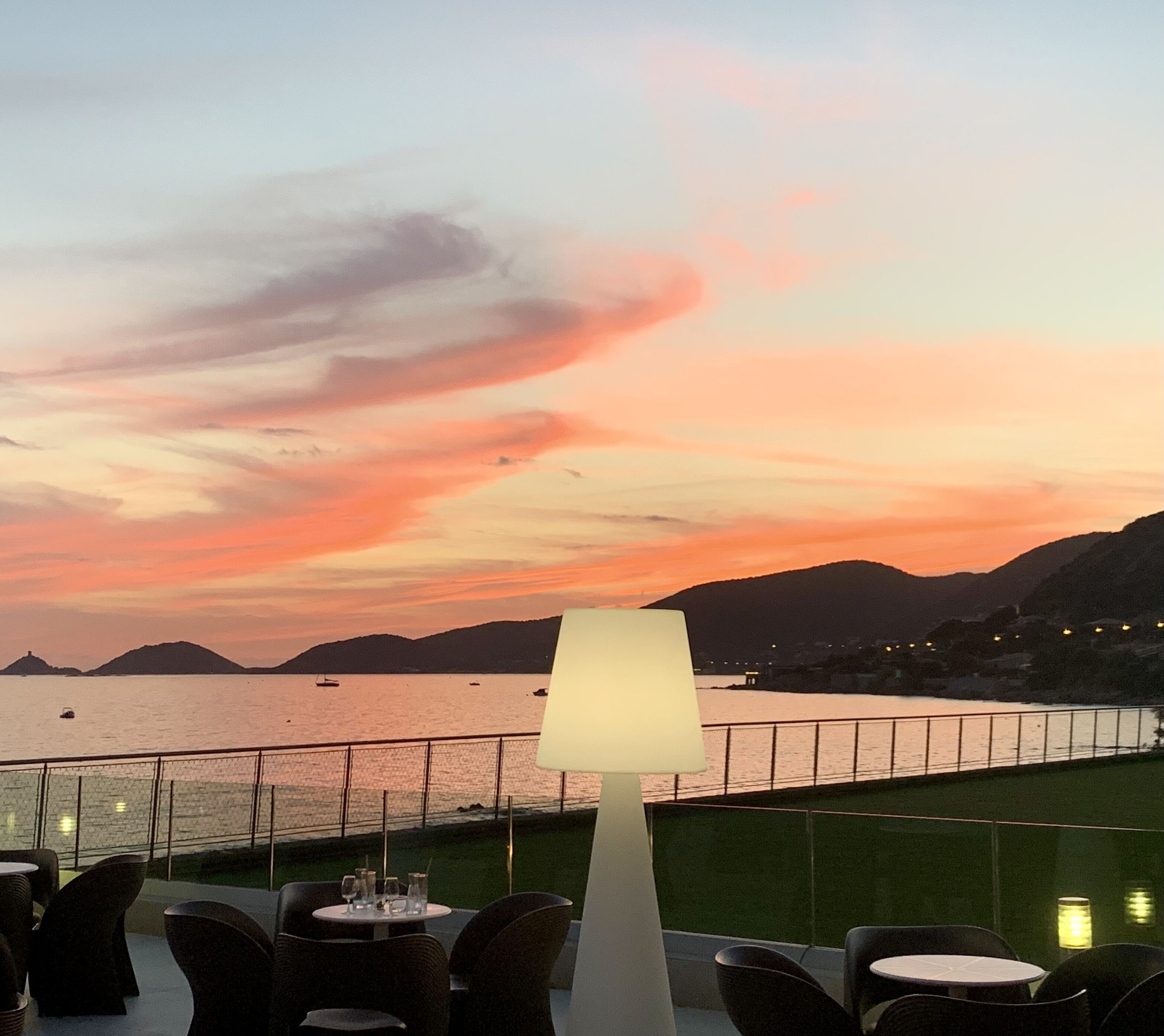 A terrace with outdoor seating overlooking a body of water and mountains during sunset.