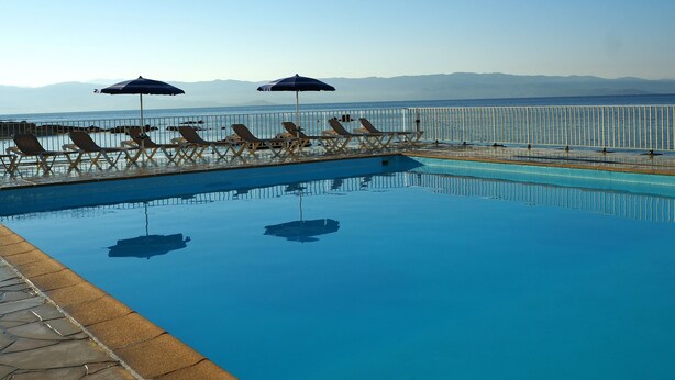 Ein Swimmingpool mit Liegestühlen und Sonnenschirmen, eine Steinumrandung und Blick auf das Meer und die Berge.
