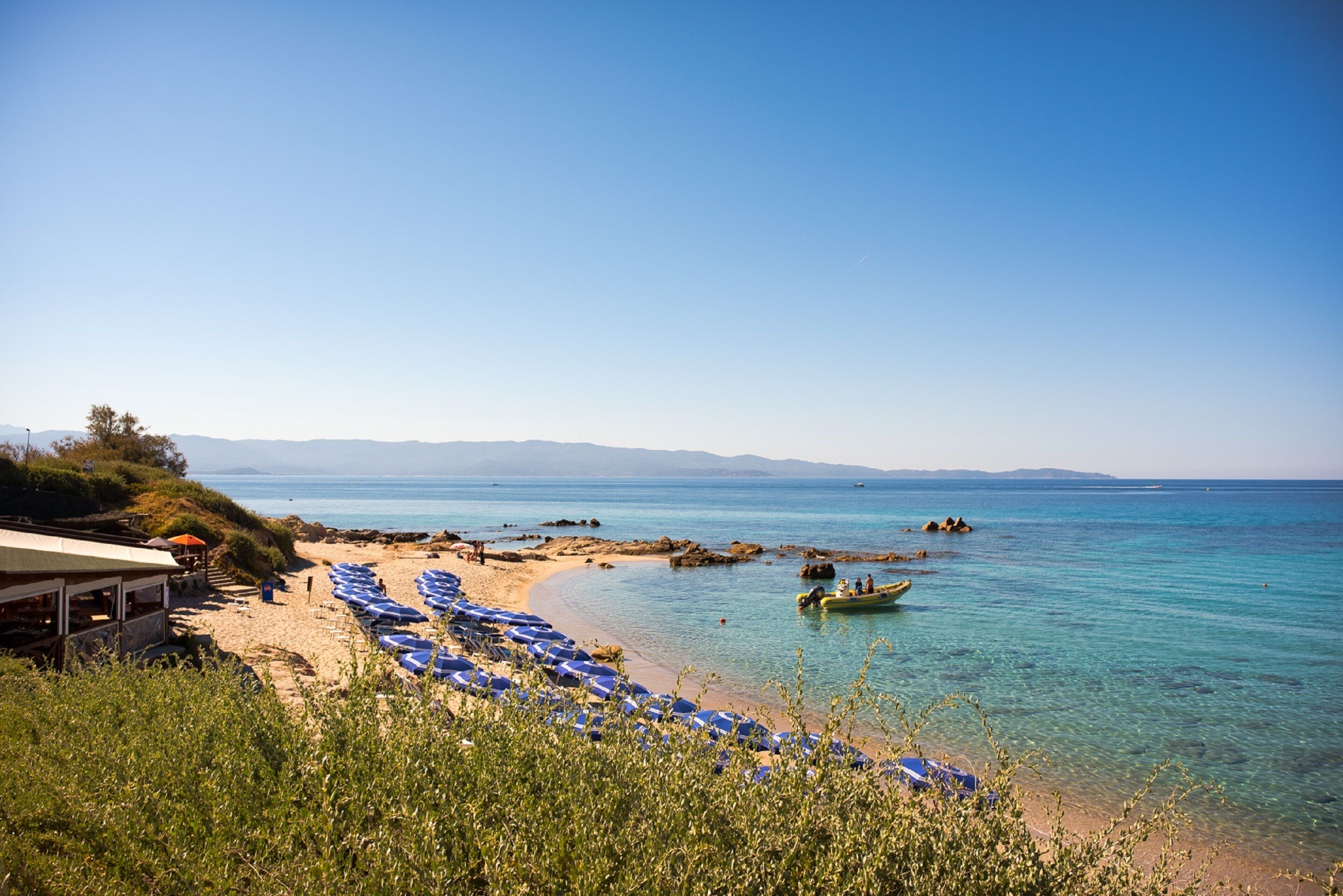 Ein Strand mit klarem, blauem Wasser, einer Reihe blauer Sonnenschirme und einem kleinen Boot.