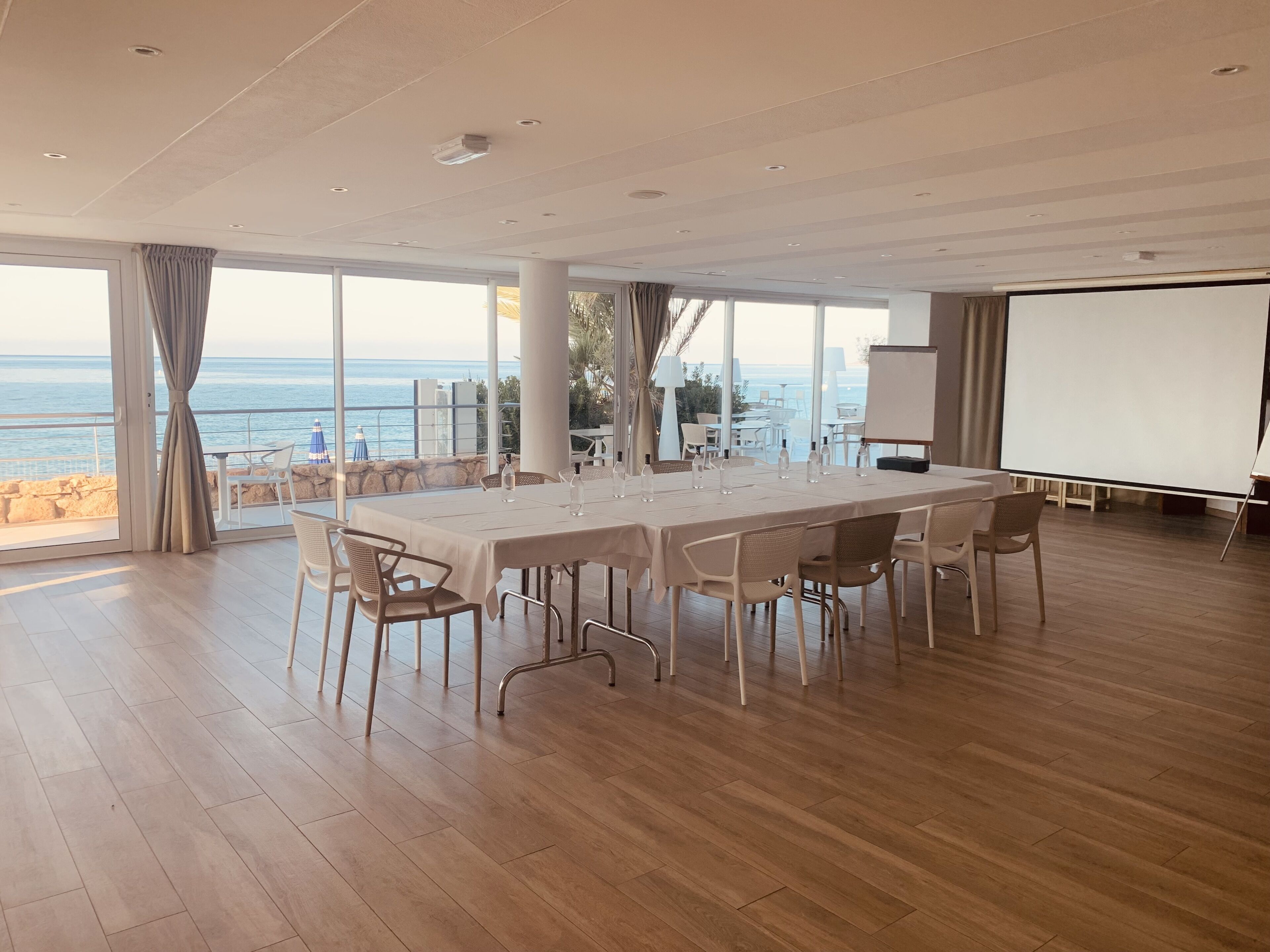 Une salle de conférence spacieuse, dotée d’une grande table et de chaises, de larges fenêtres offrant une vue sur la mer et d’un écran de projection.