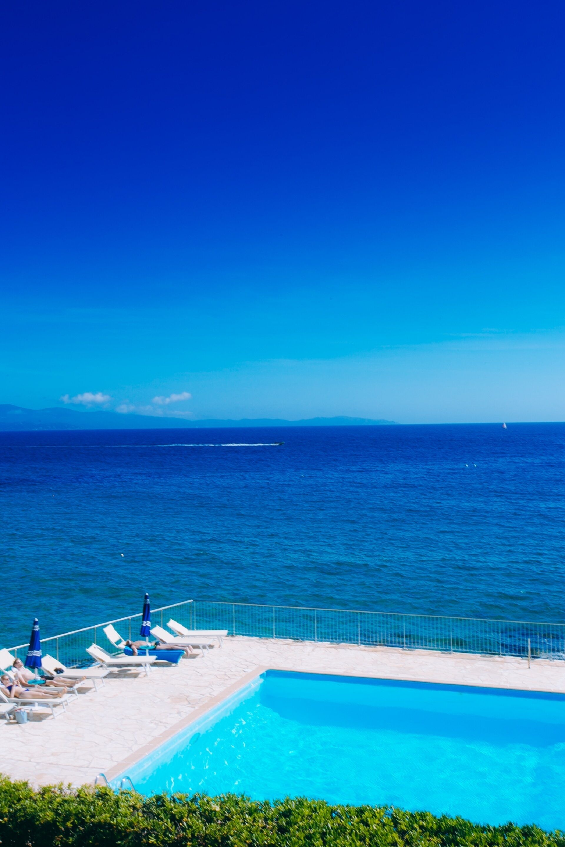 A poolside area with lounge chairs and umbrellas, overlooking a clear blue sea and distant mountains.