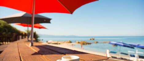 Une table en bois avec une tasse et une soucoupe blanches, un parapluie rouge et une vue sur la plage et la mer.