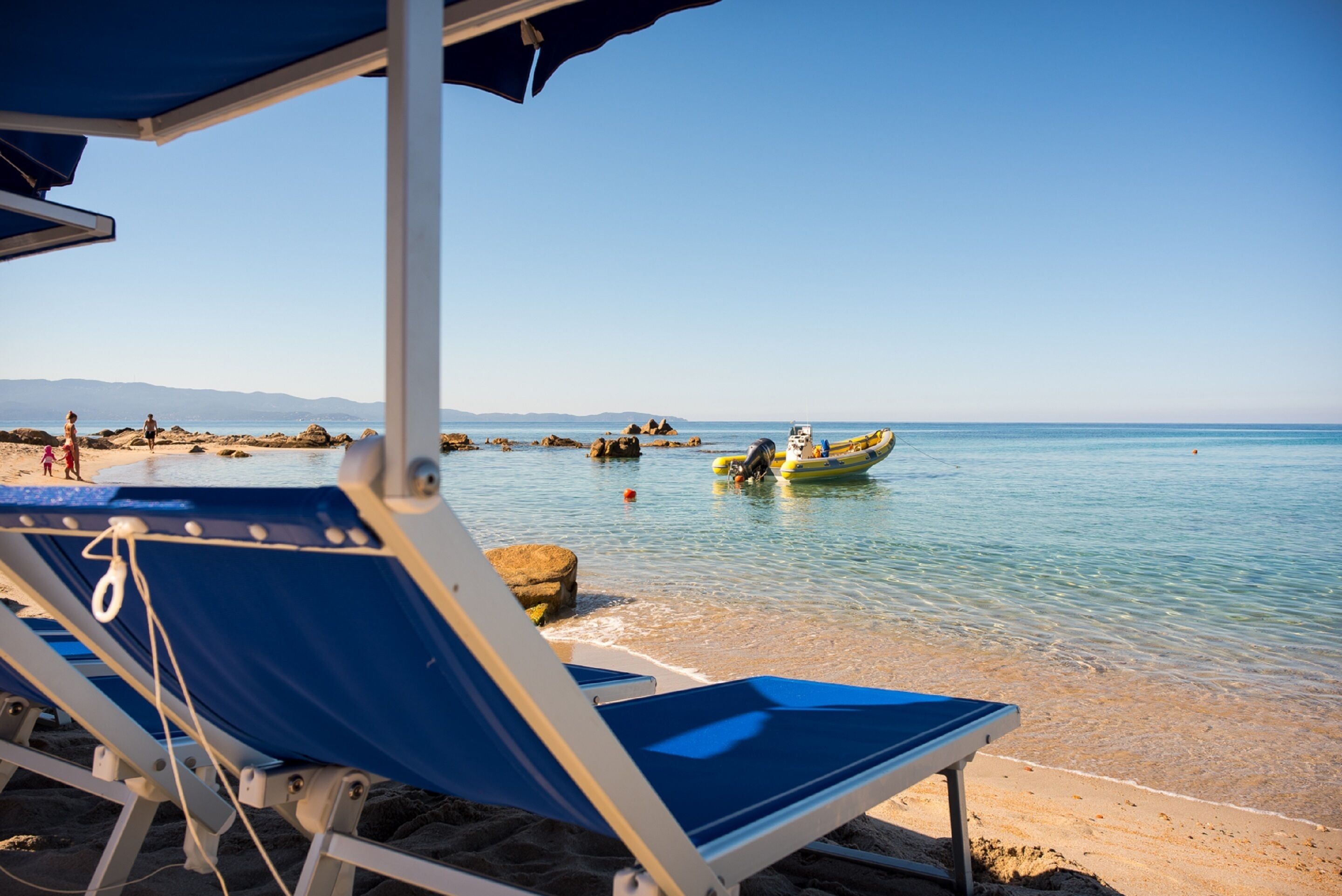 A beach with clear blue water, a row of blue beach chairs, and a small boat in the distance.