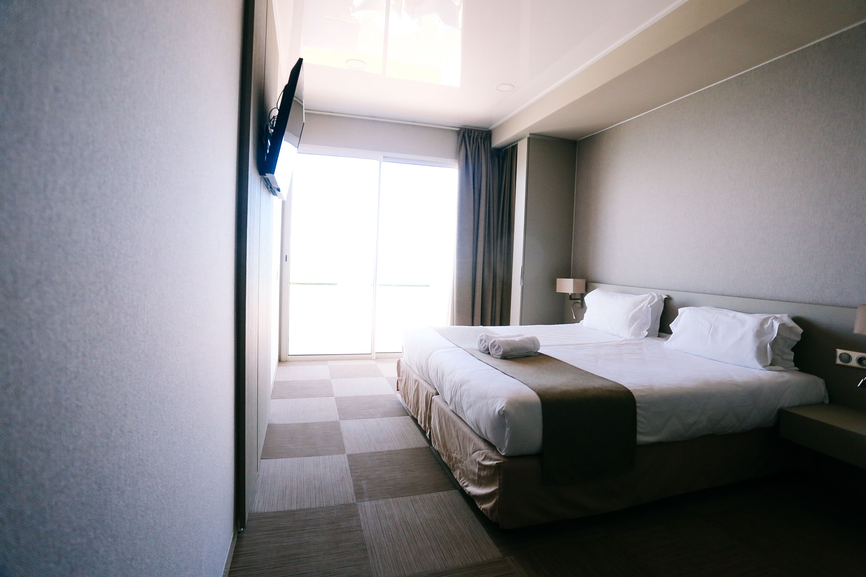 A hotel room with a large bed, a flat-screen TV mounted on the wall, a bedside table with a lamp, and a view of the outdoors through a large window.