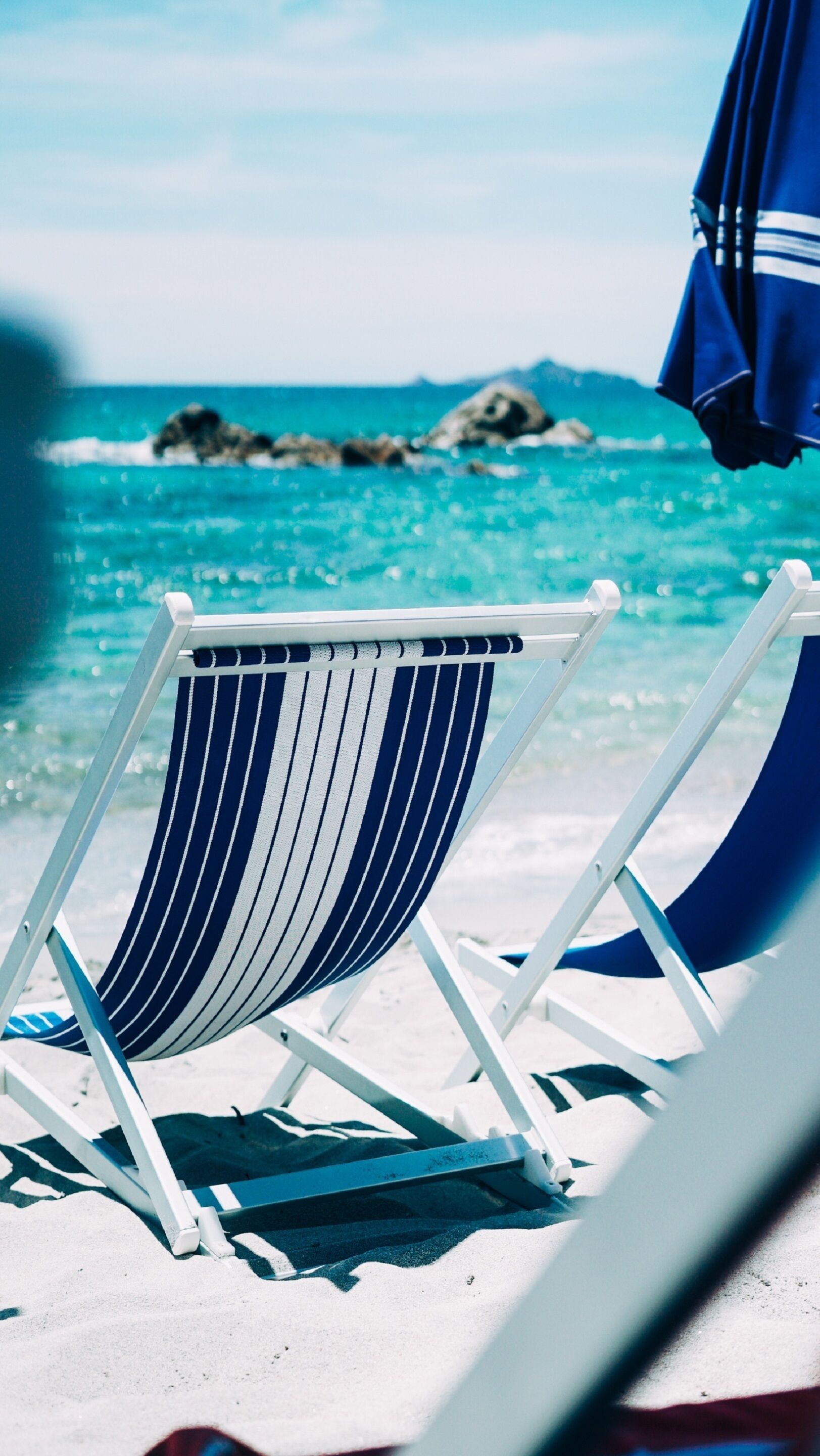 A beach chair with a striped cushion, positioned on a sandy beach with a view of the ocean and rocks in the background.