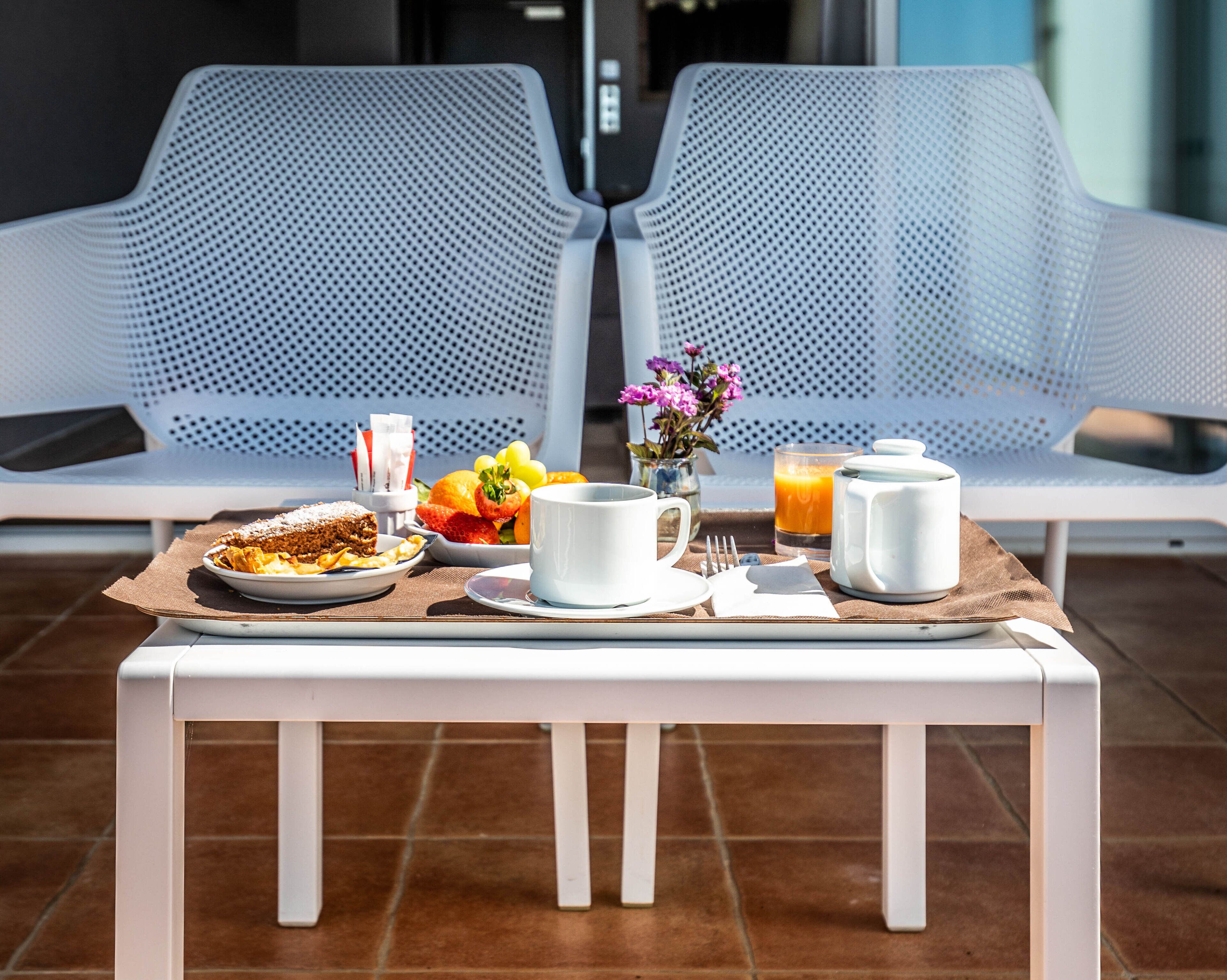 Ein Frühstücksbrett mit Speisen, einer Tasse, einem Glas Orangensaft und einem Teekännchen auf einem Tisch vor zwei weißen Stühlen mit Mesh-Geflecht.