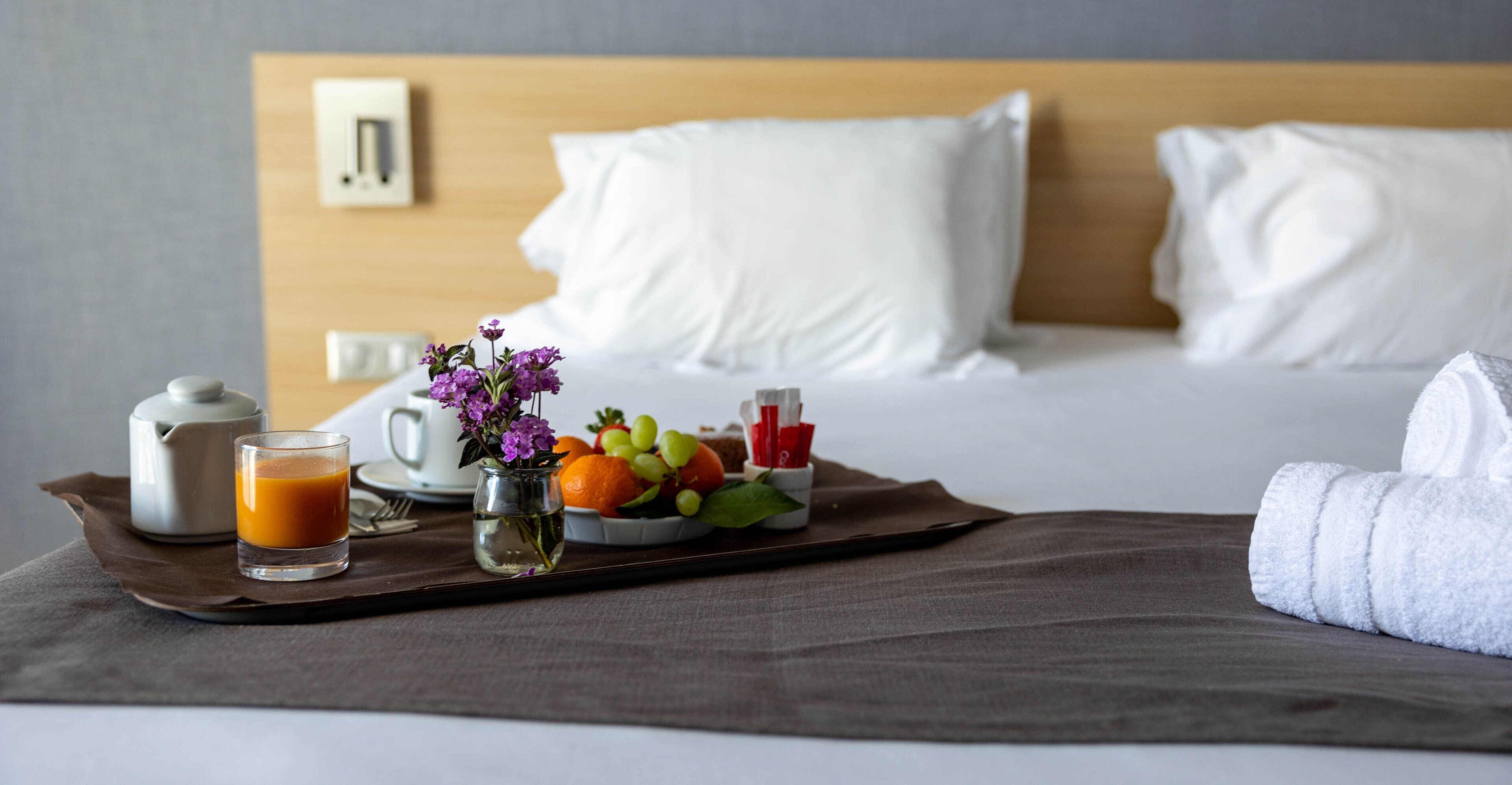 Una bandeja de desayuno ordenada con tetera, vaso de jugo de naranja, florero con flores y una fuente de fruta sobre una cama con ropa blanca y cabecera de madera.