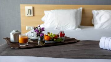 Una bandeja de desayuno ordenada con tetera, vaso de jugo de naranja, florero con flores y una fuente de fruta sobre una cama con ropa blanca y cabecera de madera.