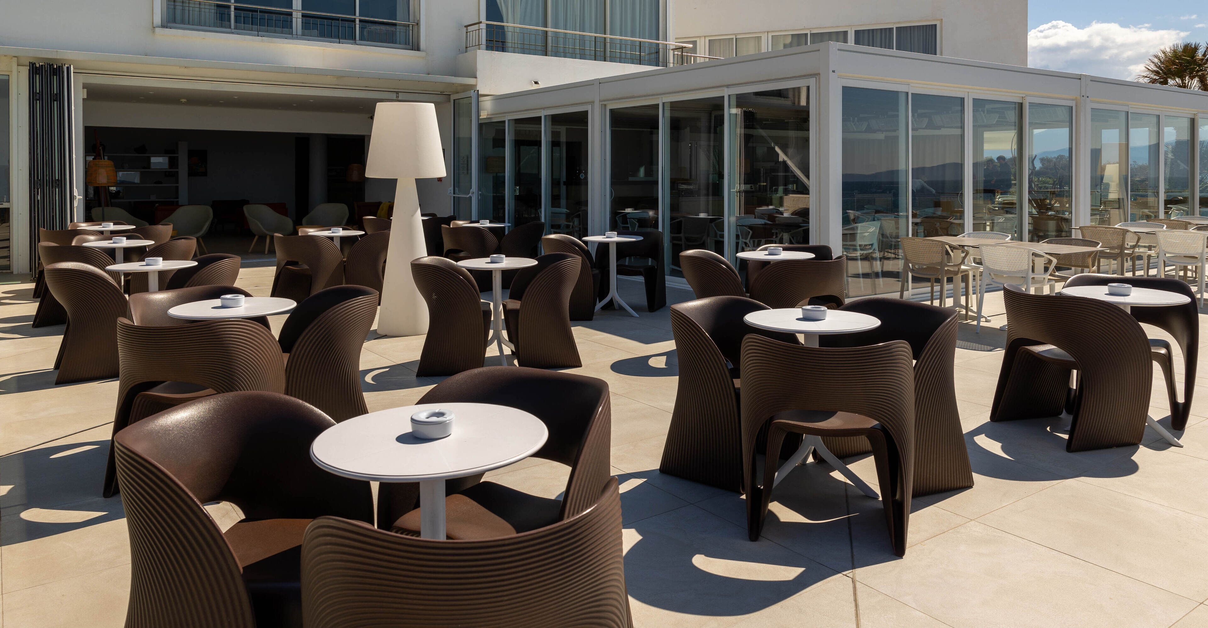An outdoor seating area with wicker chairs and round tables, large windows, and a view of the landscape.