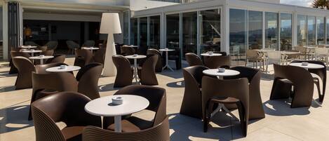An outdoor seating area with wicker chairs and round tables, large windows, and a view of the landscape.
