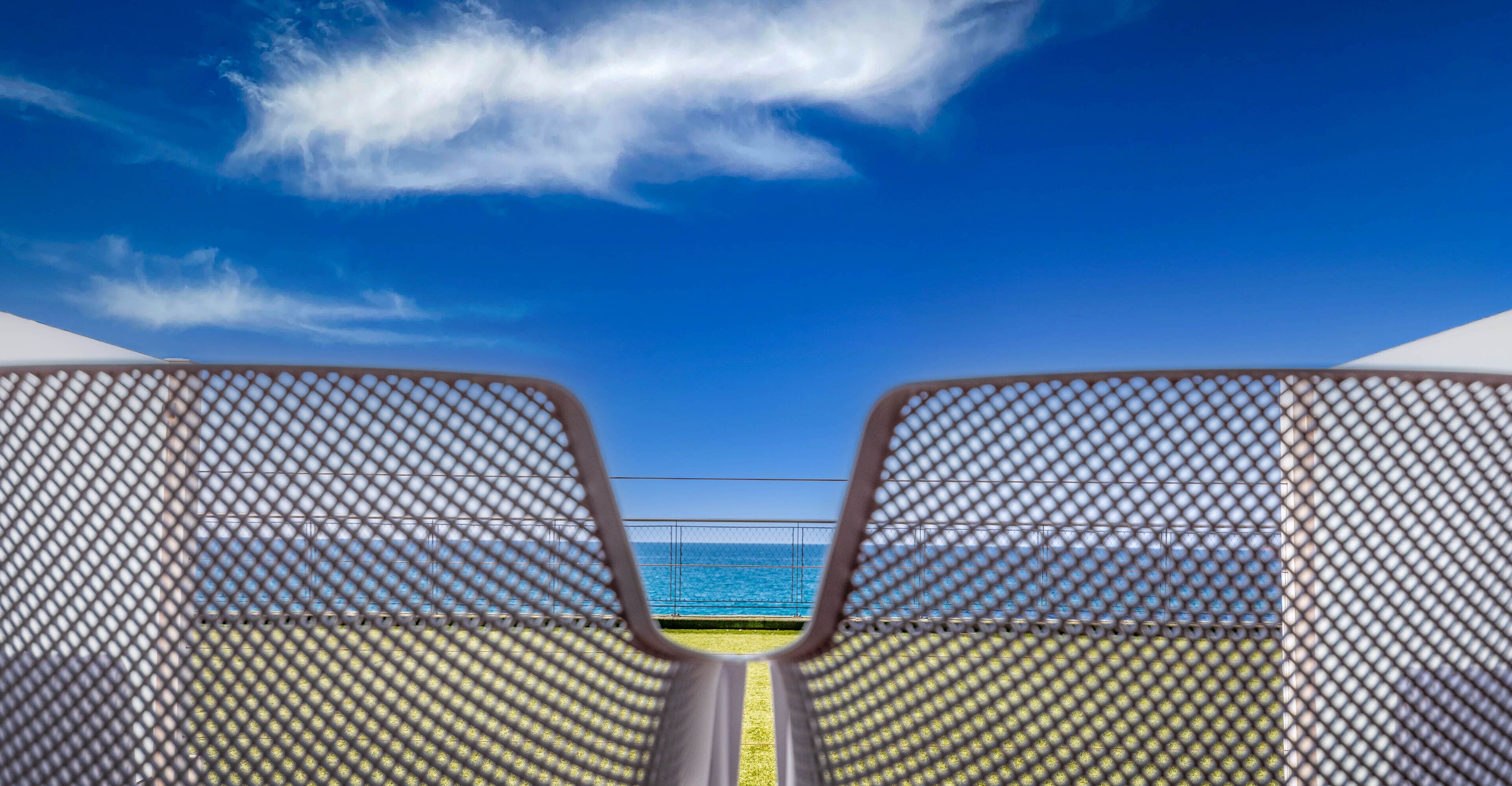 A view from a balcony with a metal railing, overlooking a clear blue sky and a calm sea.