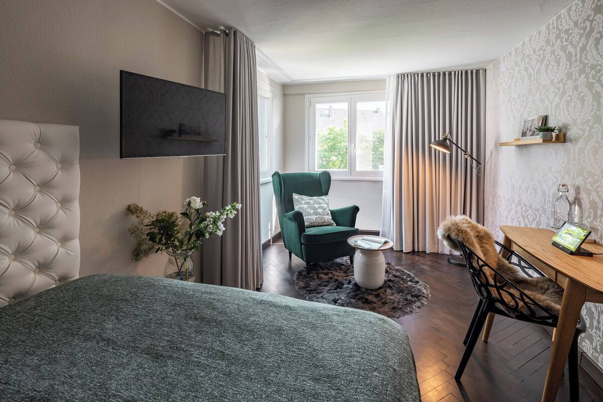 A modern hotel room with a tufted headboard, a green armchair, a wooden dining table, and a flat-screen TV mounted on the wall.