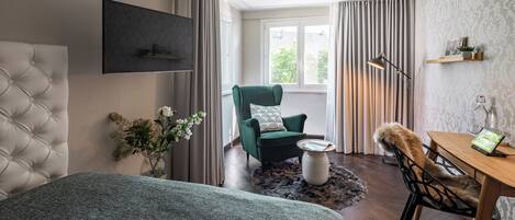A modern hotel room with a tufted headboard, a green armchair, a wooden dining table, and a flat-screen TV mounted on the wall.
