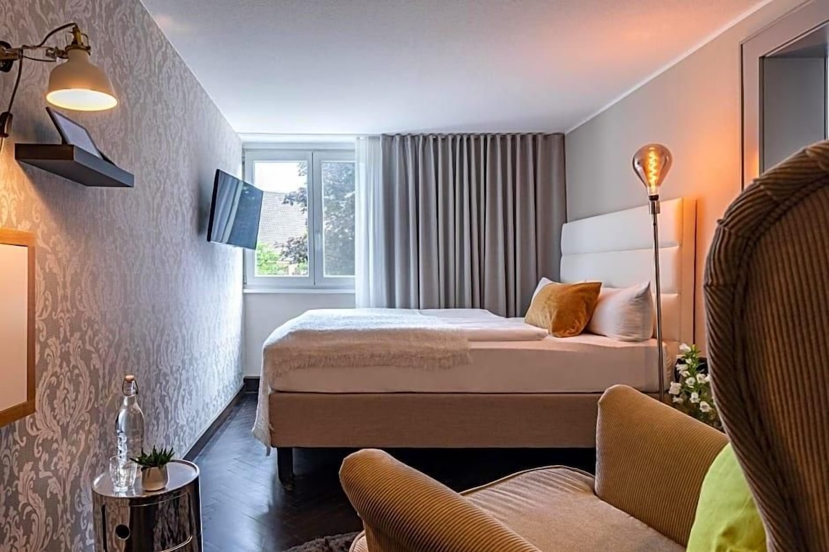 A hotel room with a bed, a sofa, a side table with a bottle, a lamp, and a TV on the wall.