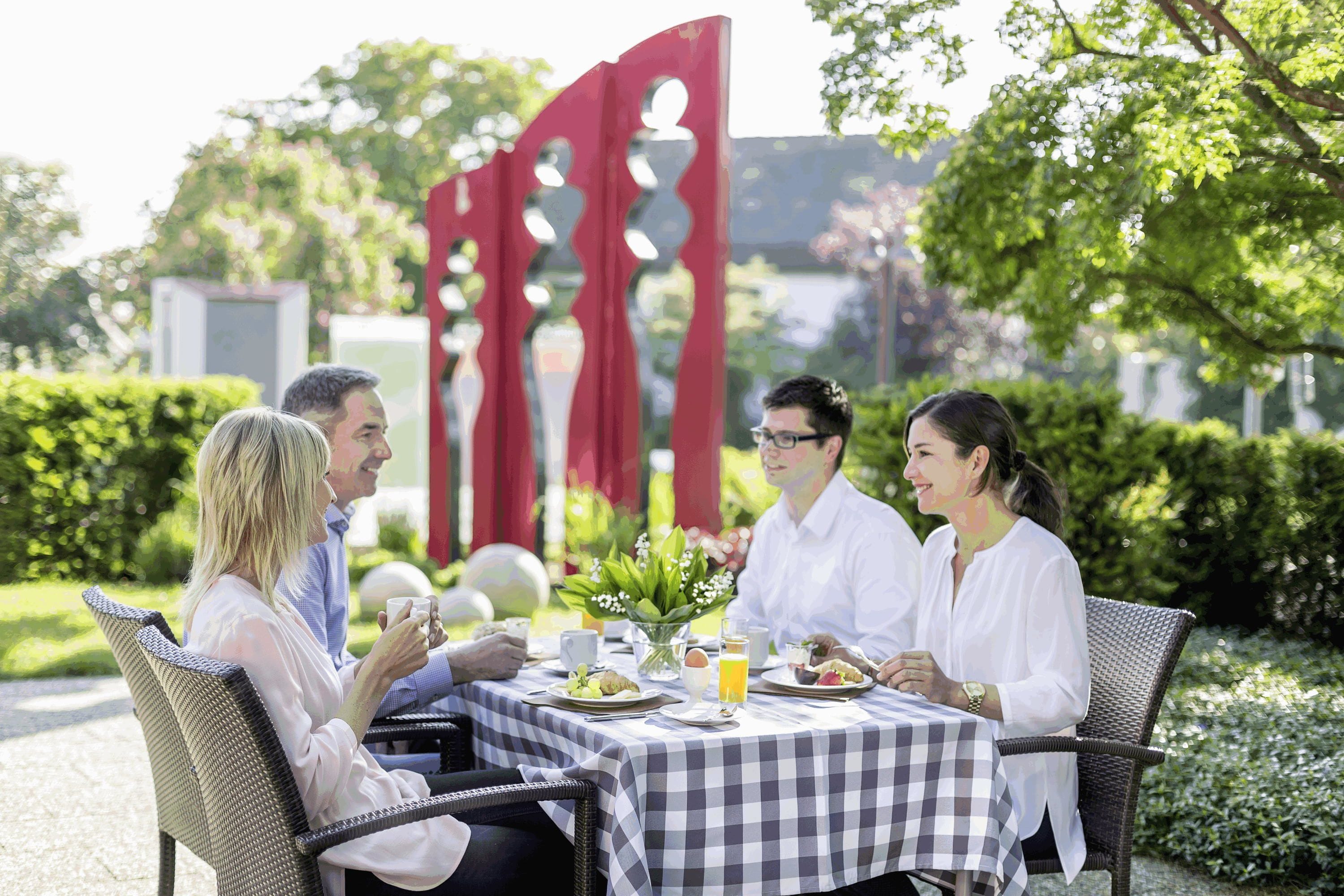 Un groupe de personnes est assis à une table à l’extérieur, où elles prennent leur repas. La table est recouverte d’une nappe à carreaux et est entourée de verdure, ainsi que d’une structure décorative rouge.