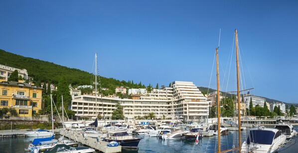 Uma marina com vários barcos atracados, um edifício grande com vários andares ao fundo e uma paisagem acidentada.
