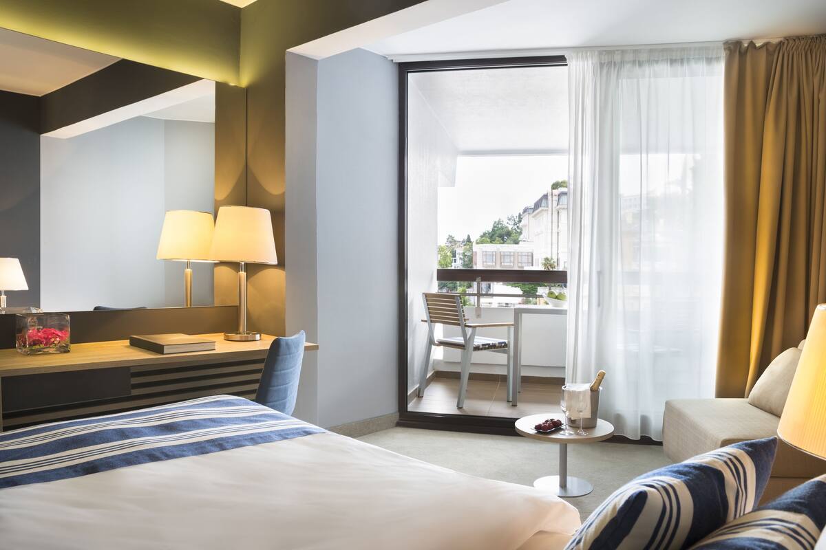 Een hotelkamer met een bed, een bureau met lampen, een kleine tafel met een fles en een glas, en uitzicht op een balkon met een stoel en een tafel.