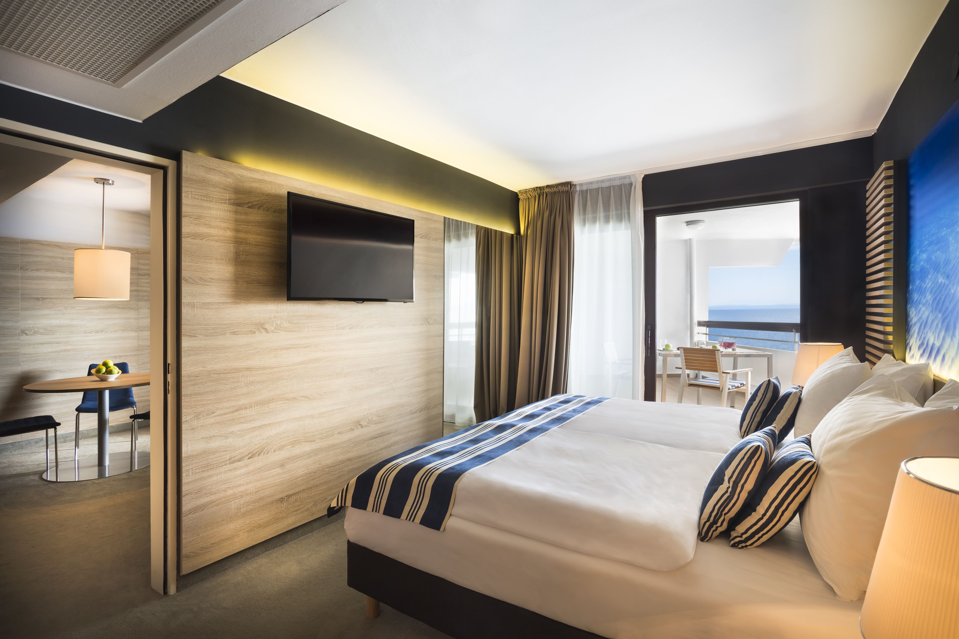 A modern hotel room with a large bed, a flat-screen TV mounted on the wall, a desk with a chair, and a view of the sea through the window.