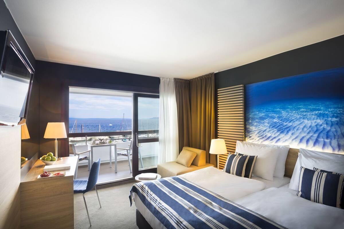 Een hotelkamer met een bed, een bureau, een stoel, een klein tafeltje, een lamp en een groot raam met uitzicht op zee.